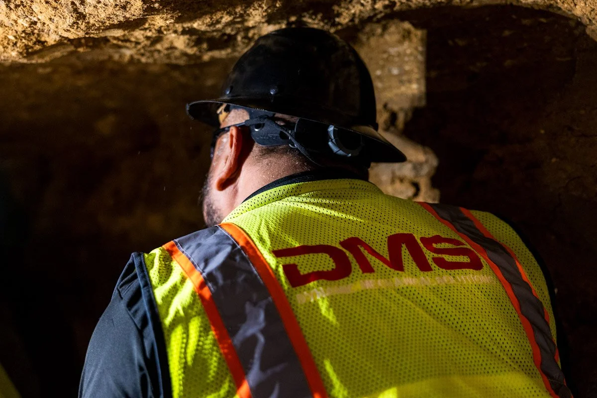 A miner wearing a yellow and black safety vest, a helmet, and glasses inside a dark underground tunnel.