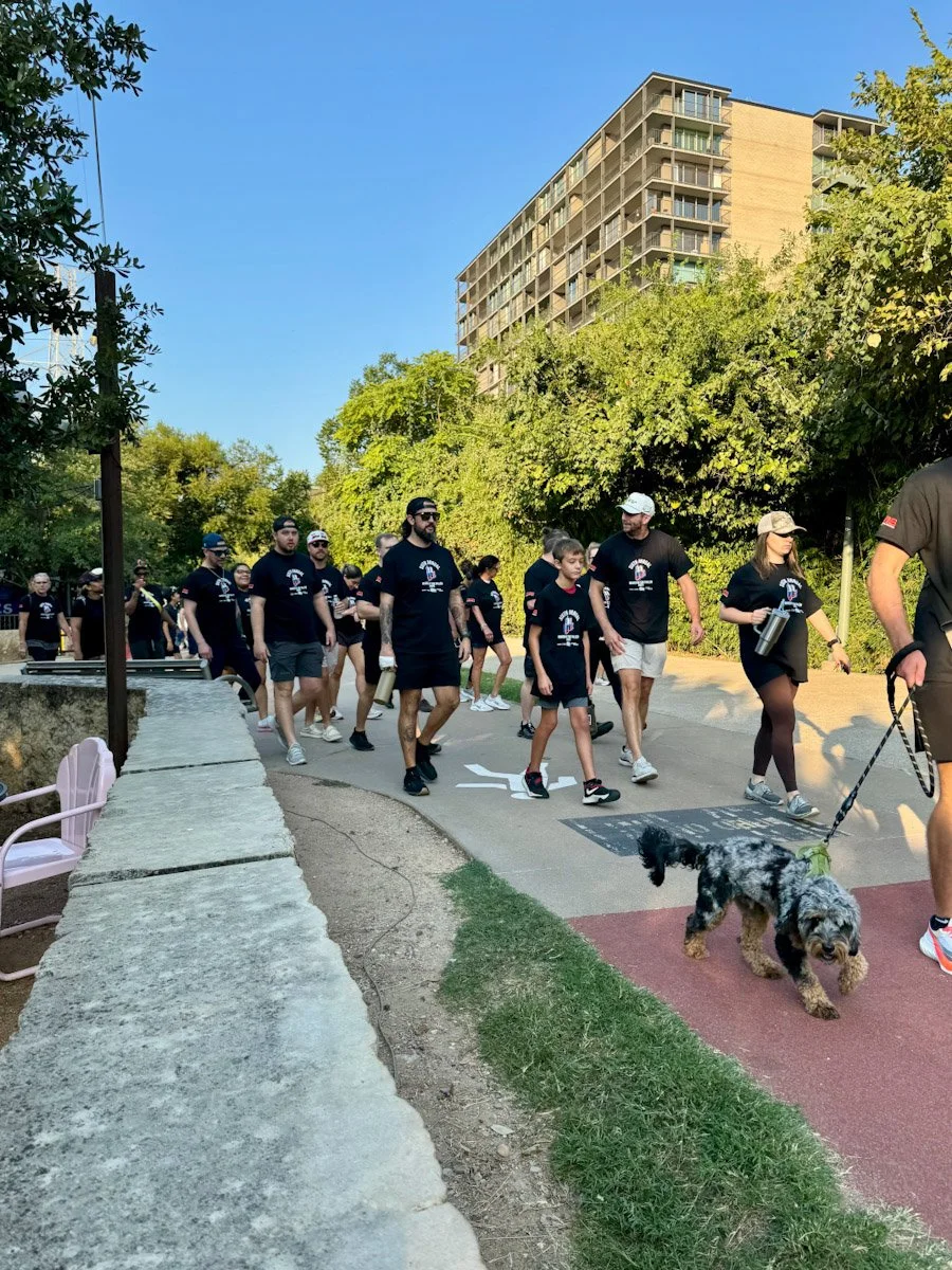 A group of people, including adults and children, walking together outdoors on a sunny day, some wearing matching black t-shirts. A woman is holding a leash with a small, fluffy dog walking on a red path. There are trees and a tall building in the background.