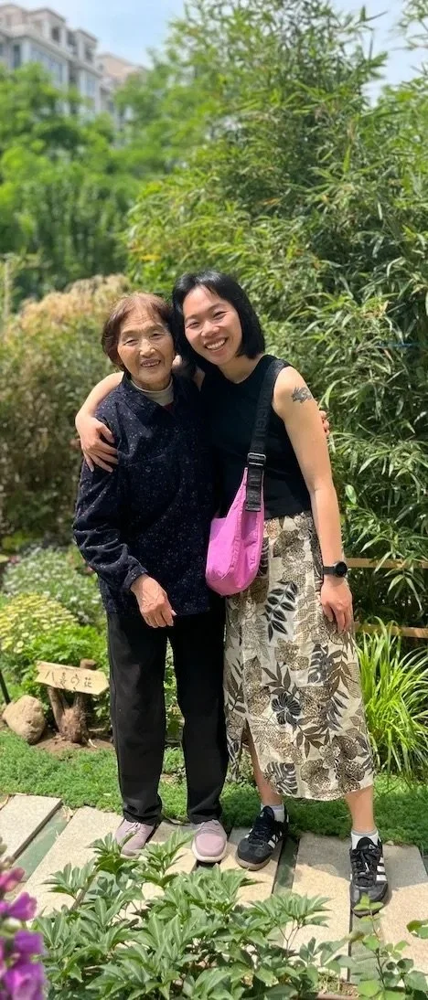 Two women smiling and hugging each other outdoors in a garden with green plants and trees.