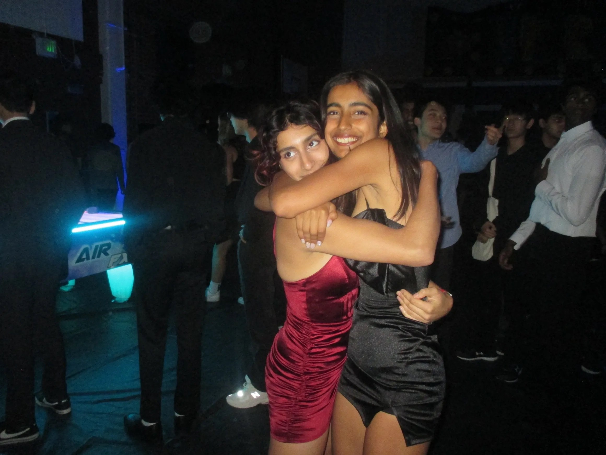 Two women hugging and smiling at a party or nightclub, with a crowd of people in the background.