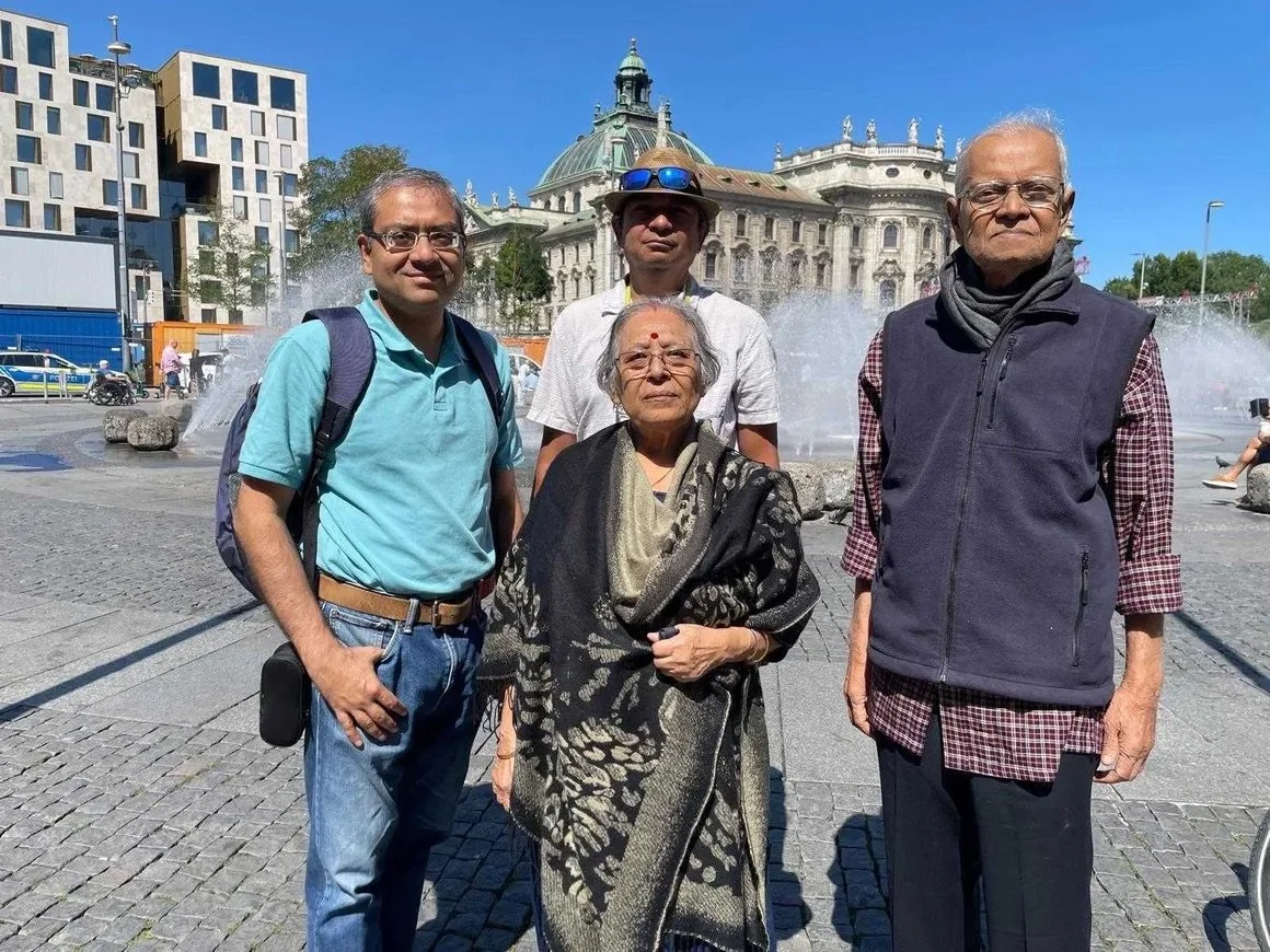 Four people posing in front of a fountain and historic building on a sunny day.