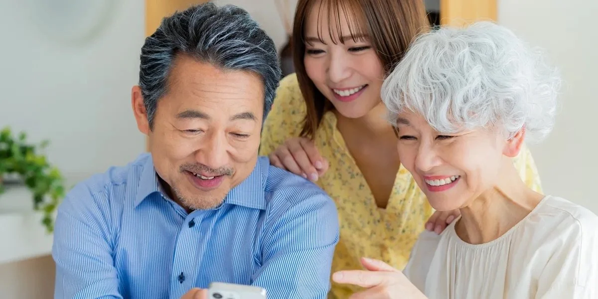 Three people smiling and looking at a smartphone together, with one older woman pointing at the phone, a middle-aged man, and a young woman.