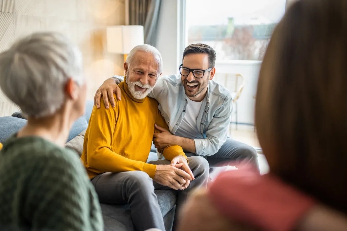 Four people, including an elderly man with white hair and a beard, are sitting on a couch and smiling, enjoying a conversation in a well-lit living room.