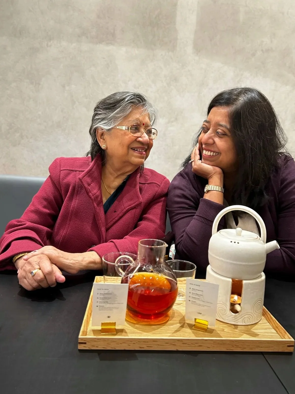 Two women, older and younger, sitting at a table with tea and a tea warmer, sharing a joyful moment and smiling at each other.