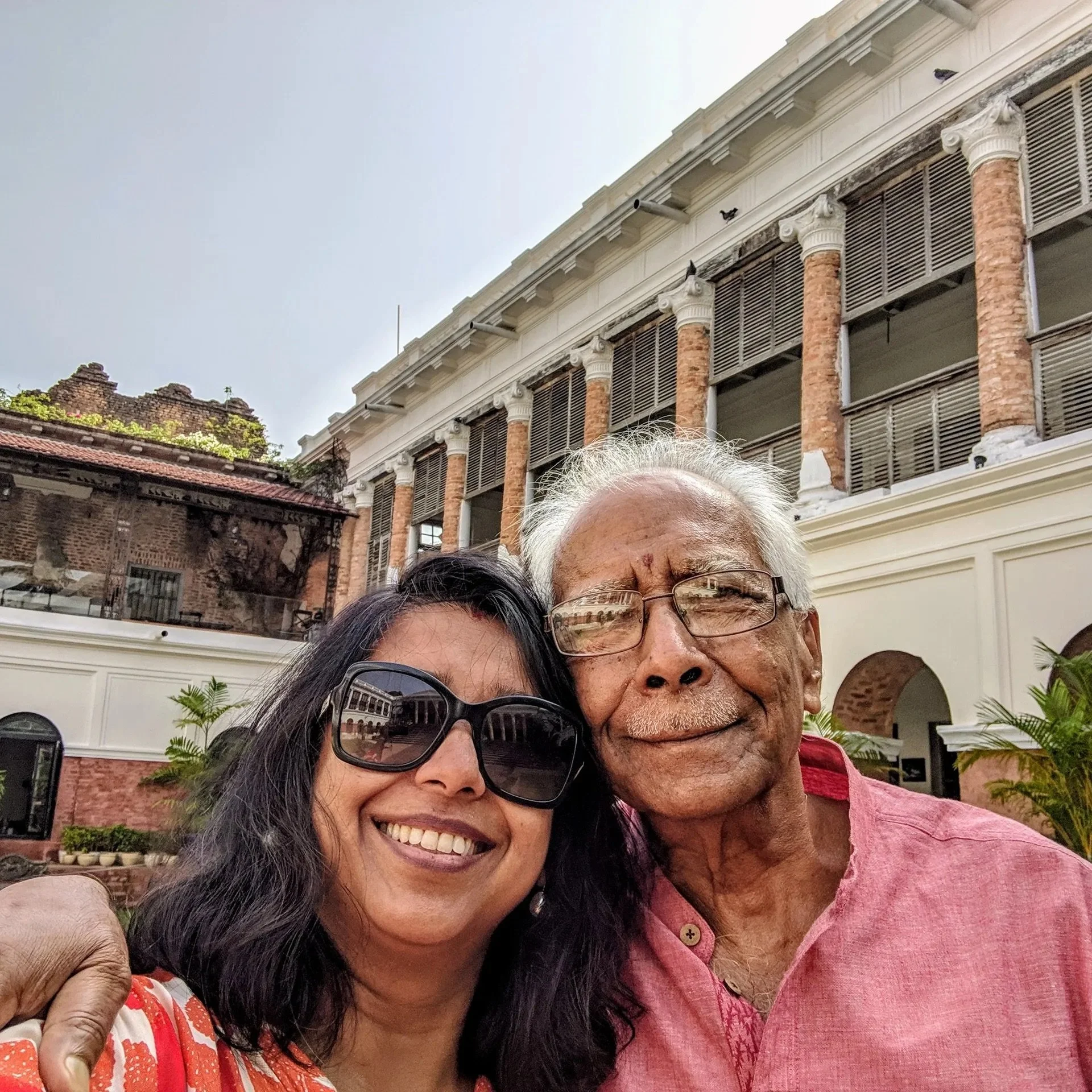 A woman and an elderly man smiling for a selfie outside a building with columns and a white and brick facade.