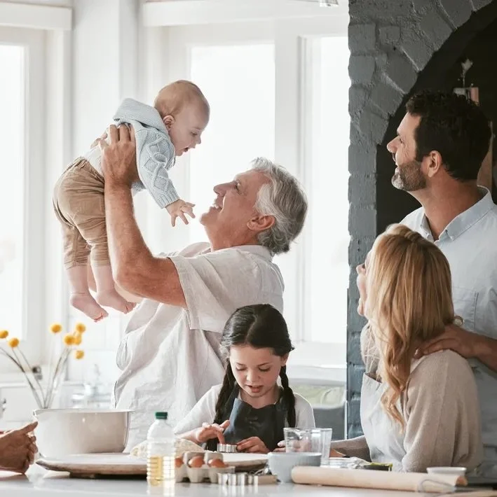 Grandfather lifting a baby in a bright kitchen while family members watch, including a man and woman, and a young girl with braided hair playing with a phone.