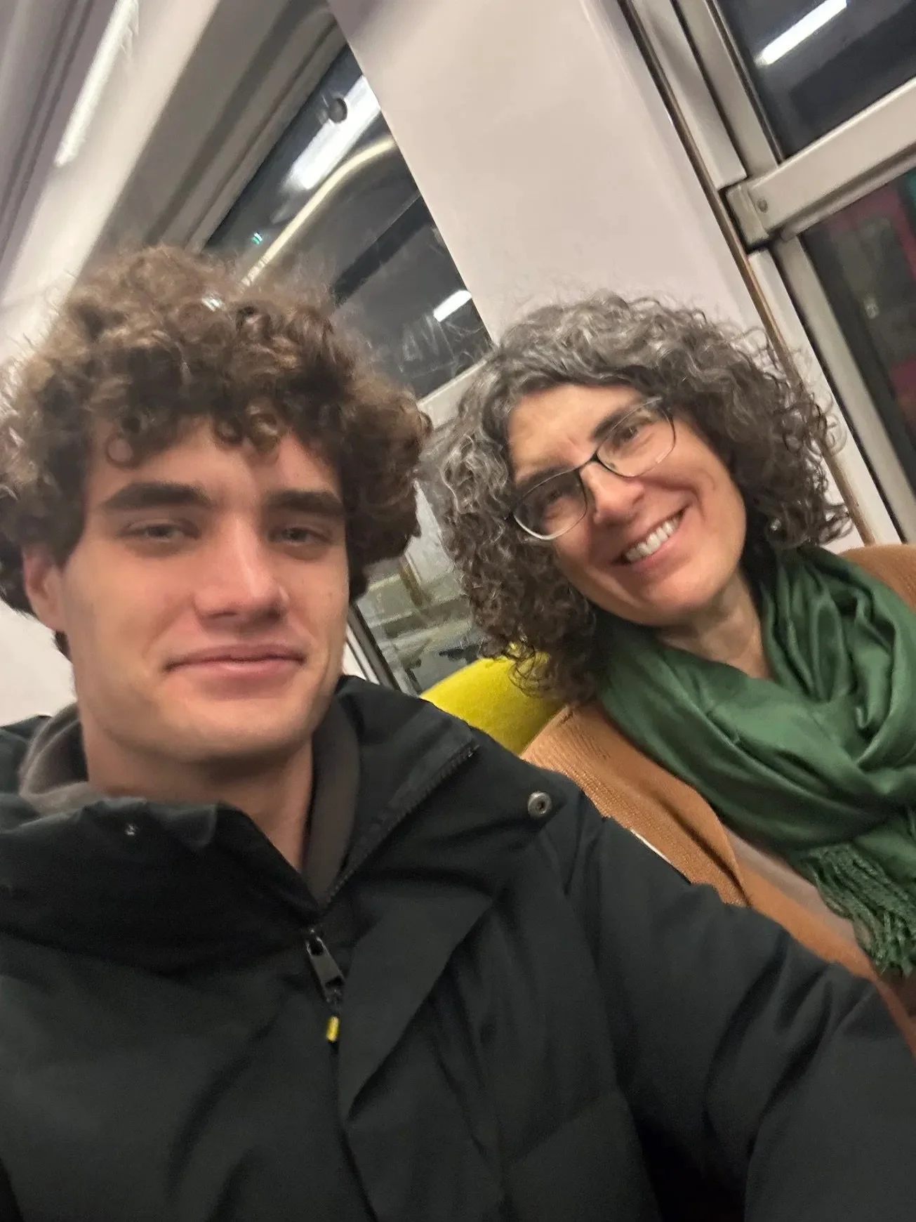 Two men sitting together on a train or bus, smiling at the camera. The person on the left has curly brown hair and is wearing a black jacket. The person on the right has curly gray hair, glasses, and is wearing a green scarf and brown jacket.