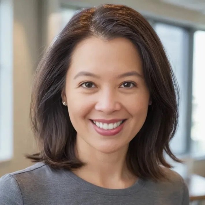 A woman with shoulder-length brown hair smiling in an indoor setting with large windows.