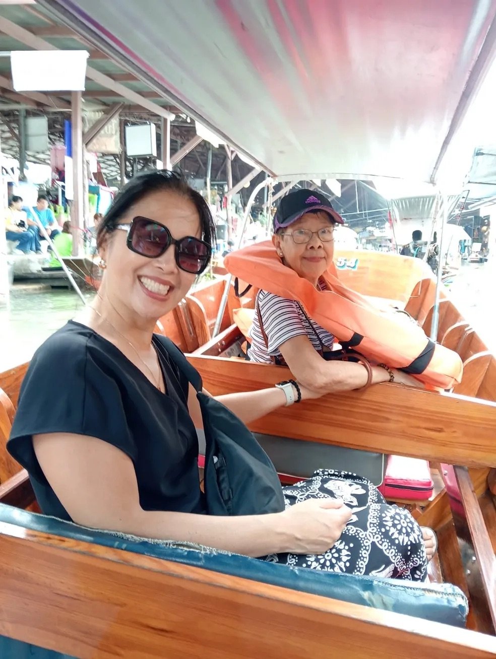 Two women sitting in a boat, smiling at the camera, with a lively outdoor market or dock scene in the background.