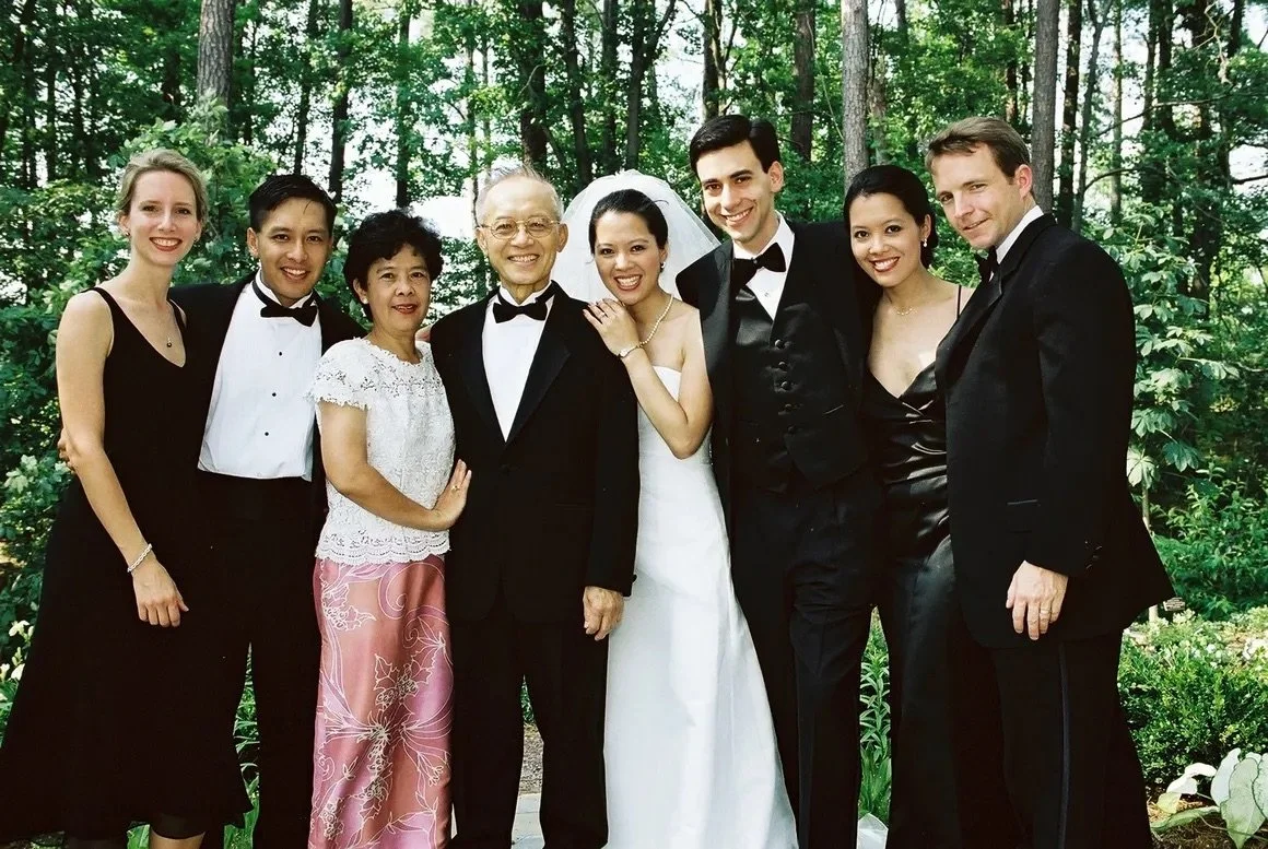 Group of people dressed in formal attire, standing outdoors in a wooded area, celebrating a wedding.