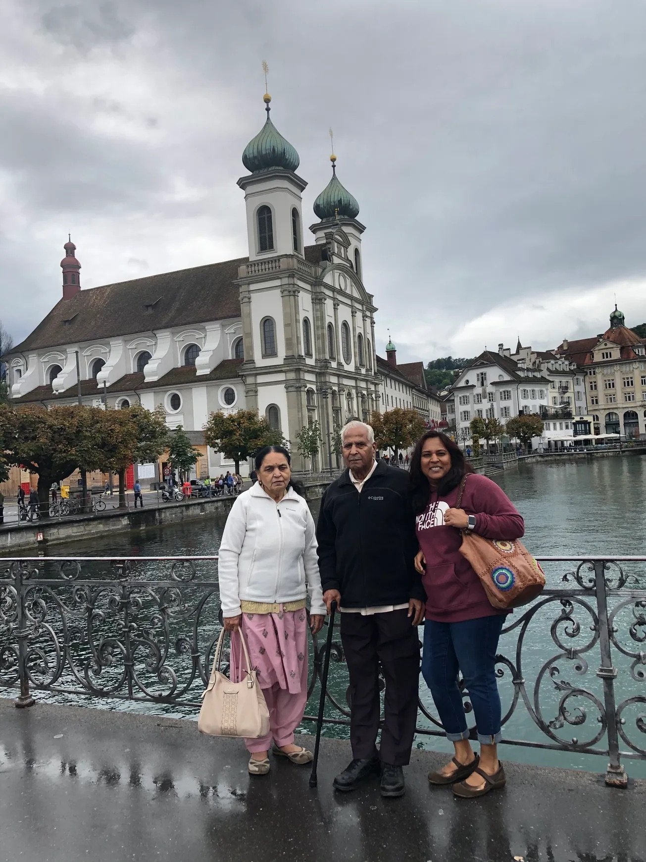 Three people standing on a bridge with a river behind them, in front of a historic church with two green domes, in a European city.
