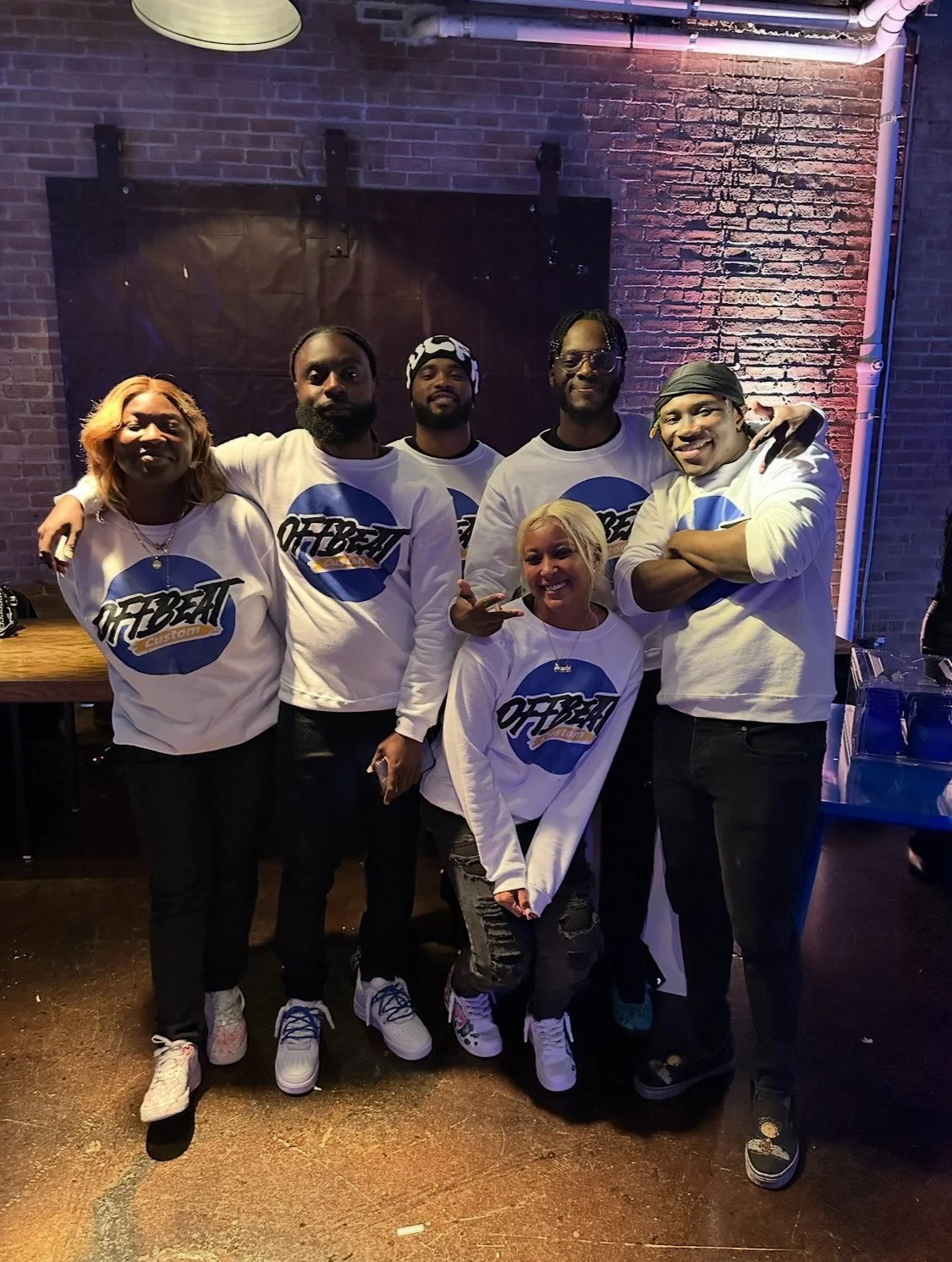 Group of six people wearing matching Offbeat shirts, standing in front of a brick wall, smiling and posing together.