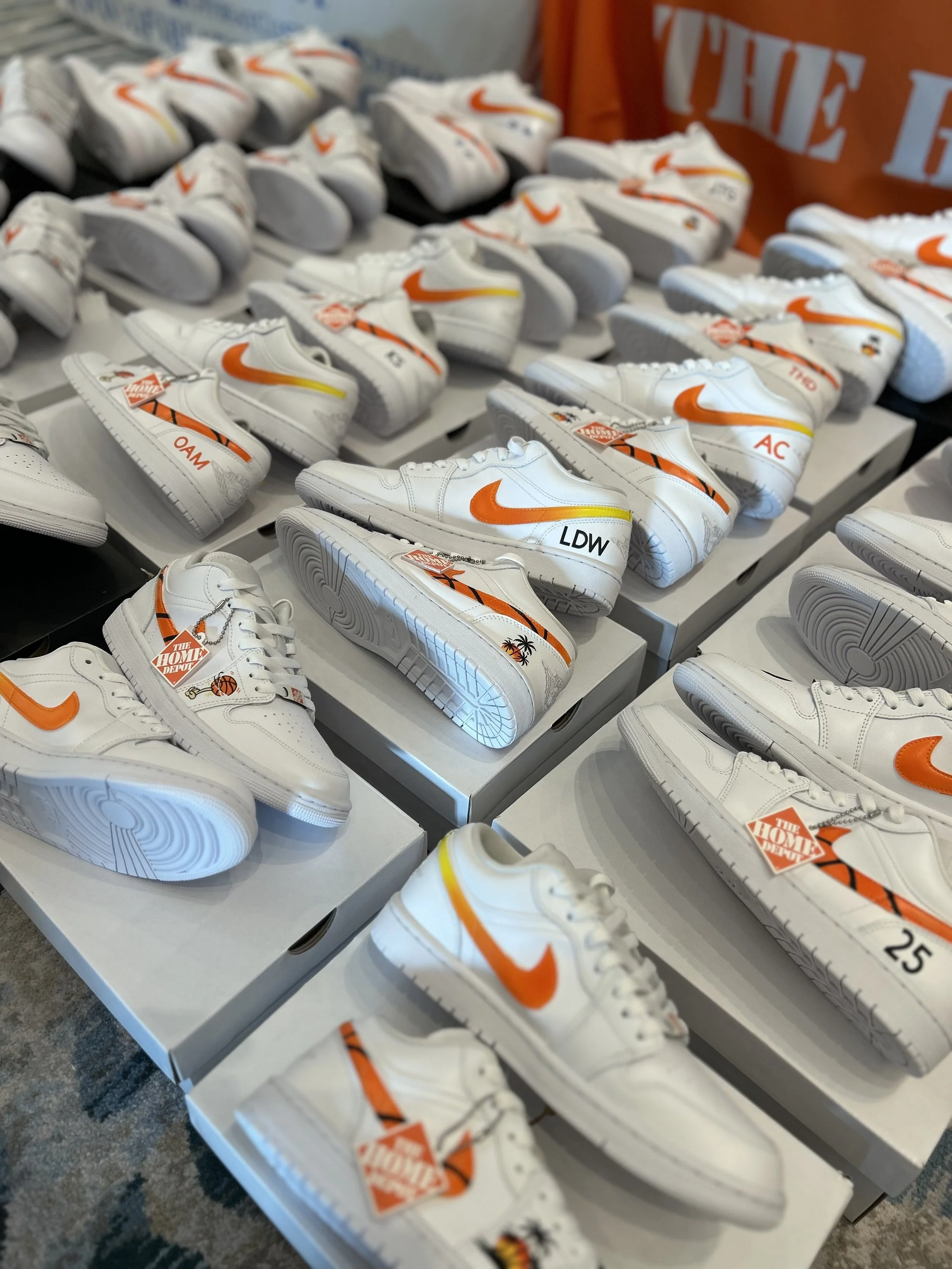 Multiple pairs of white Nike sneakers with orange swoosh logos and orange and black stripes, displayed on white shoeboxes at a store or exhibition.