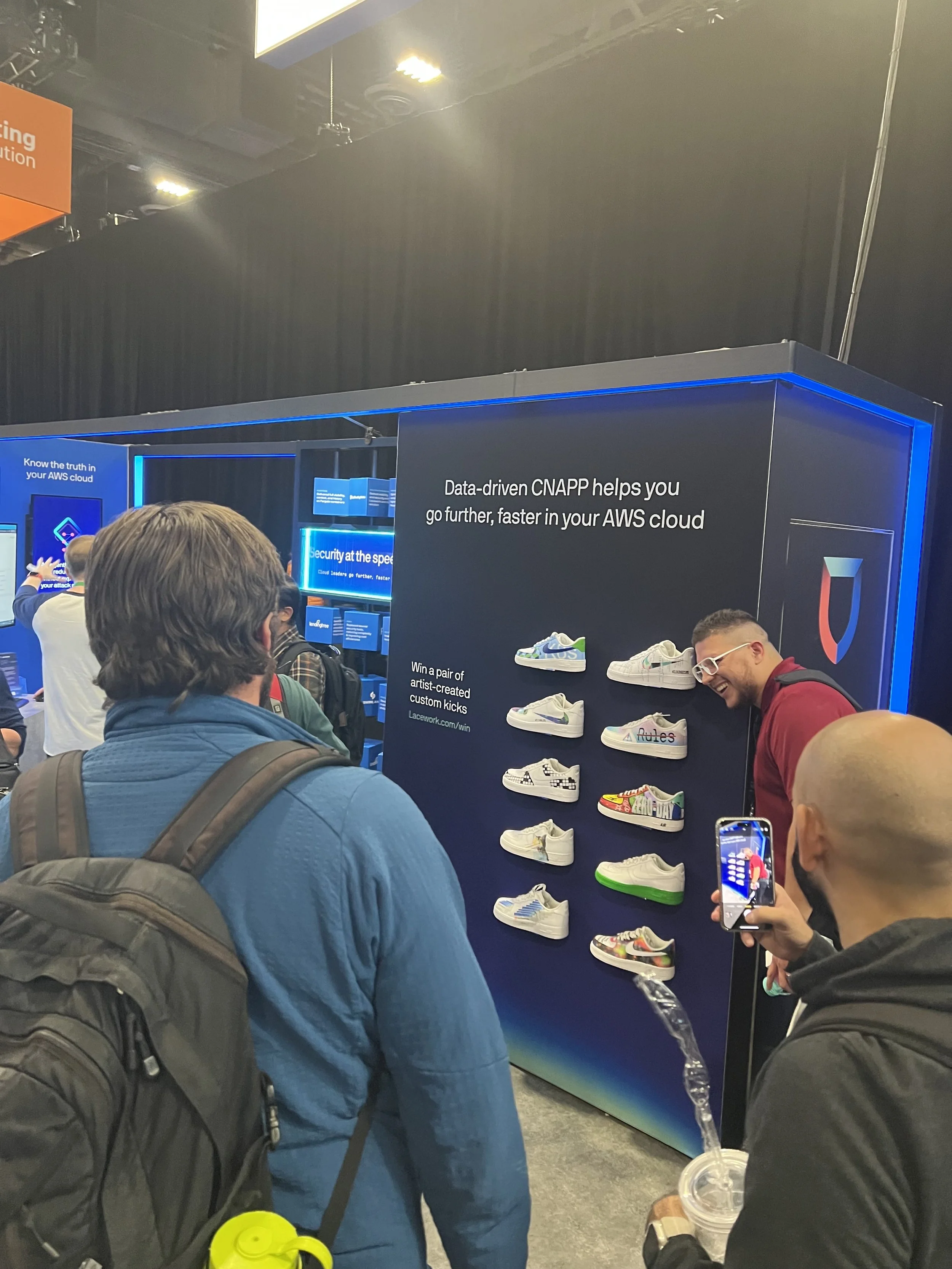 People gathered around a display at a tech event, with shoes on a board and a sign mentioning data-driven CNAPP helping with AWS cloud, and a contest for custom artist-created kicks.