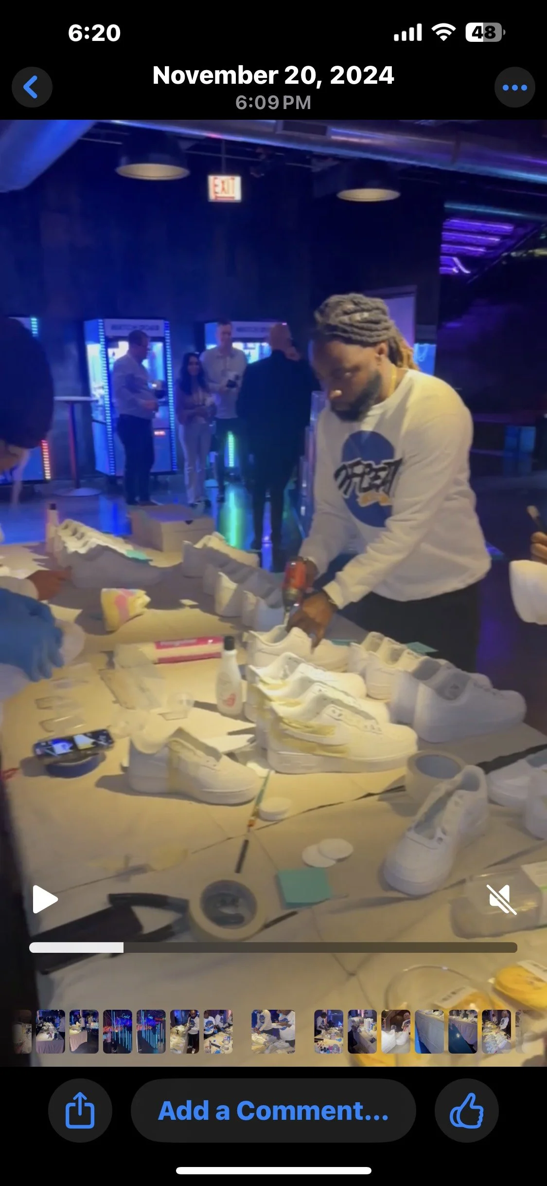Man with gray dreadlocks wearing a white sweatshirt working on white sneakers at a table during a sneaker-themed event with people in the background and blue neon lighting.
