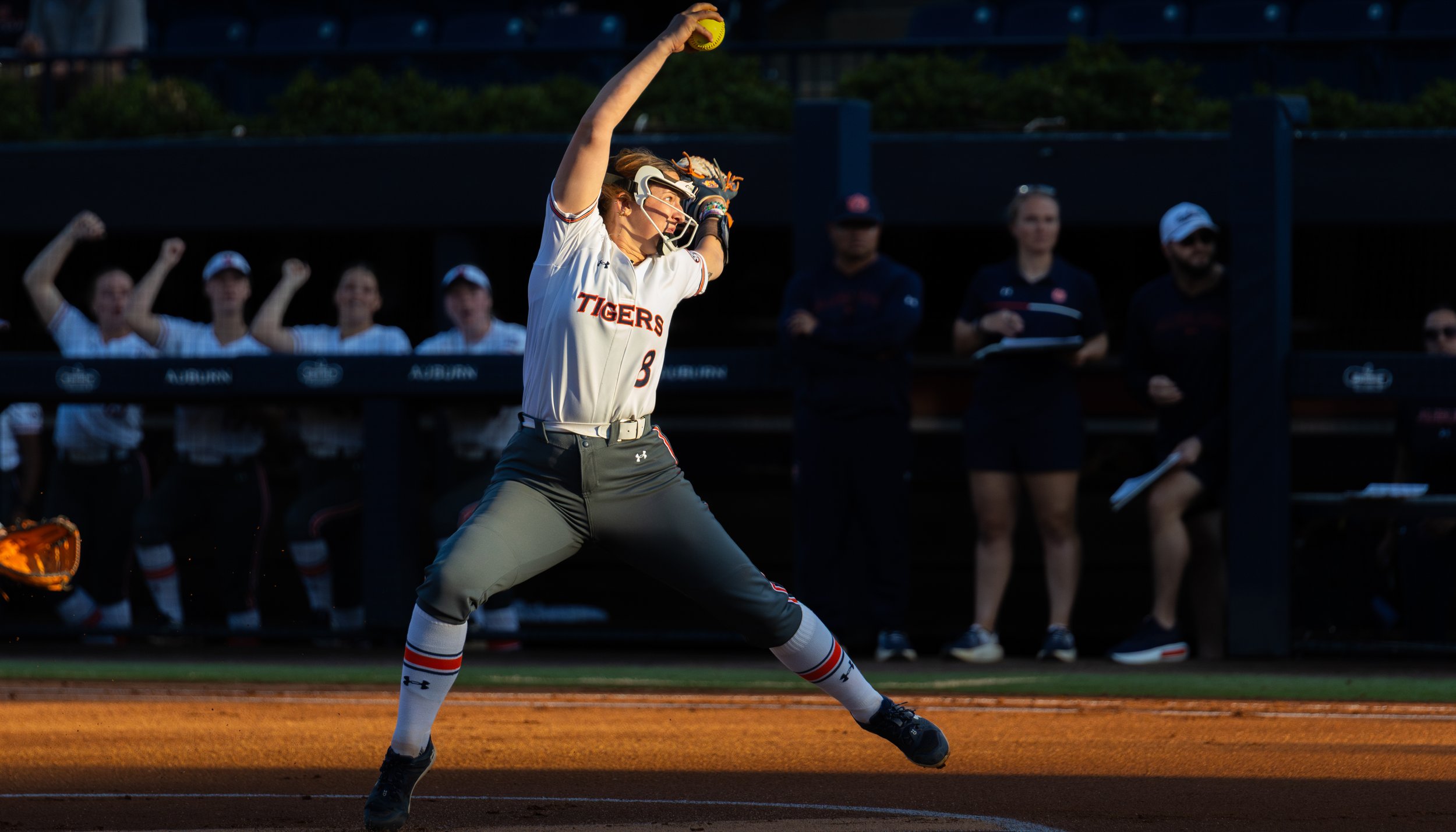 A female softball player in a white uniform with 'TIGERS' written on it, holding a yellow softball above her head, preparing to pitch on the field.