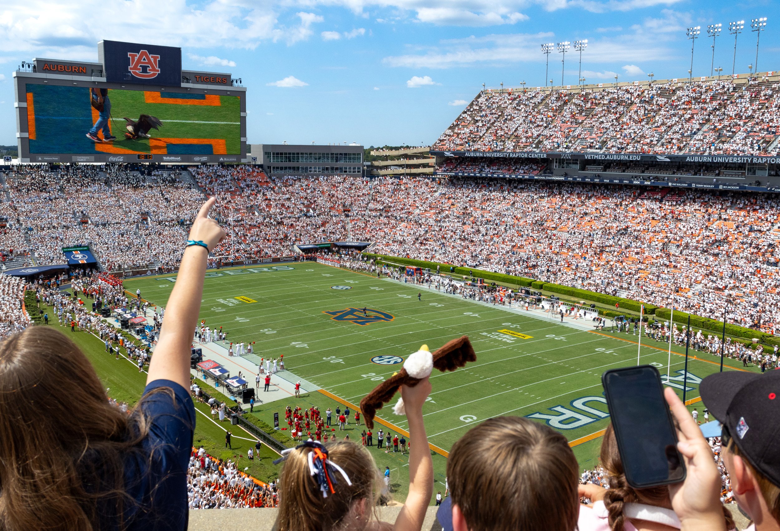 A football stadium filled with fans, including children, watching a game. The field has the Auburn University logo, and the scoreboard displays a Eagle on a football field.