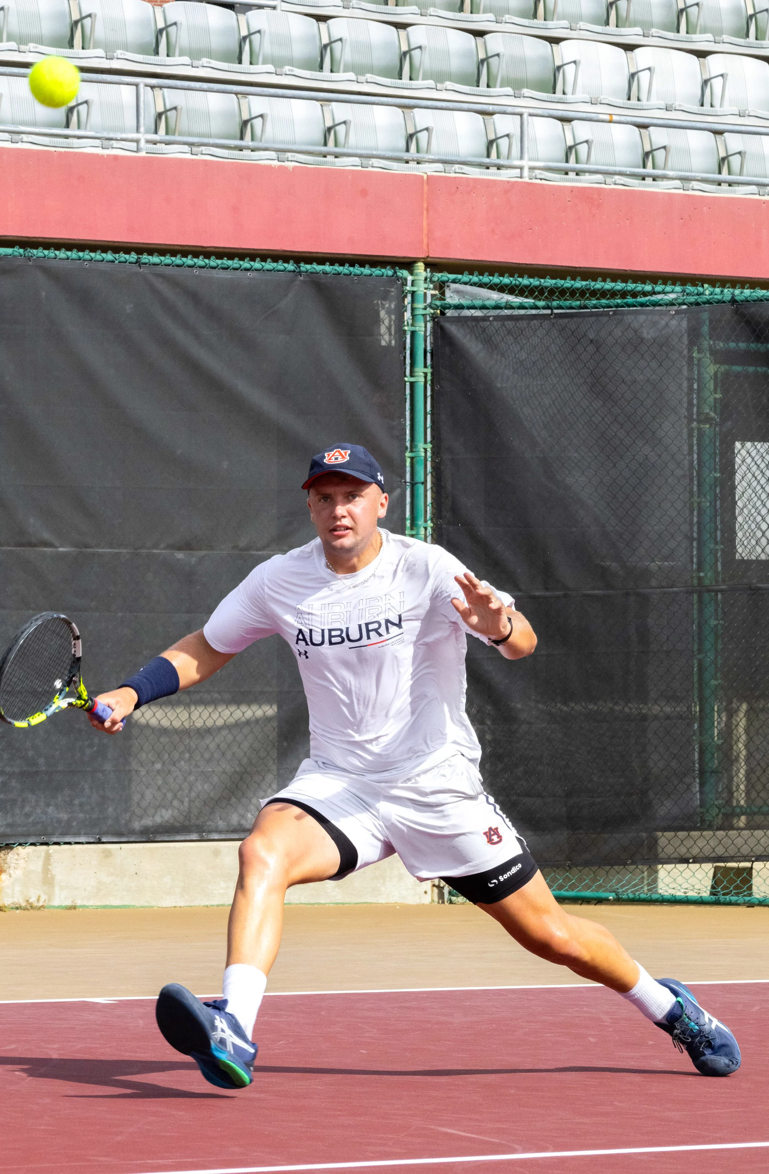 A male tennis player wearing a white Auburn shirt and shorts, holding a tennis racket, is lunging on a tennis court to hit a bright yellow tennis ball coming toward him.