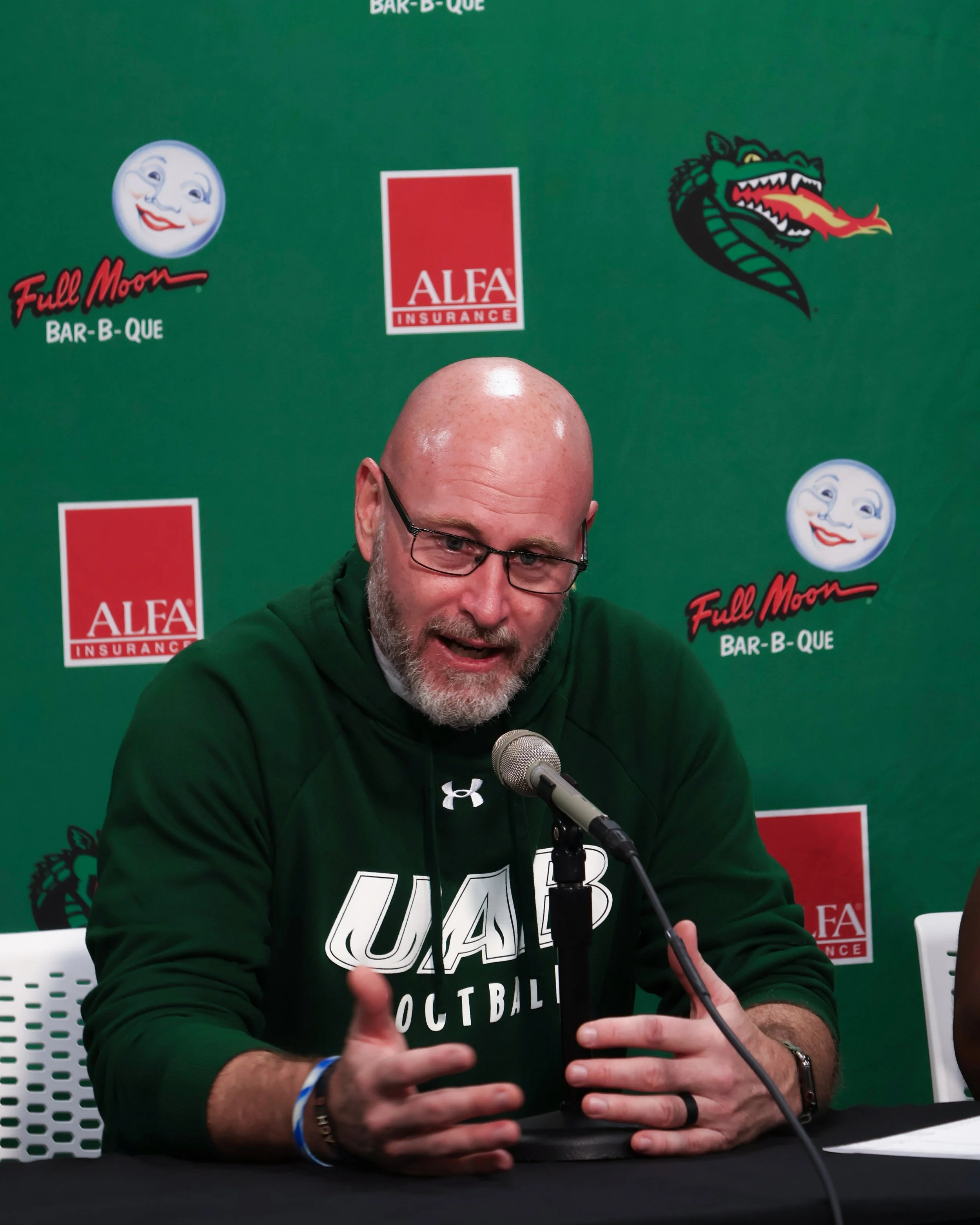 A man with glasses and a beard, wearing a black UAB basketball hoodie, speaking into a microphone at a press conference with a green background featuring logos of Full Moon Bar-B-Que, Alfa Insurance, and a UAB's mascot.