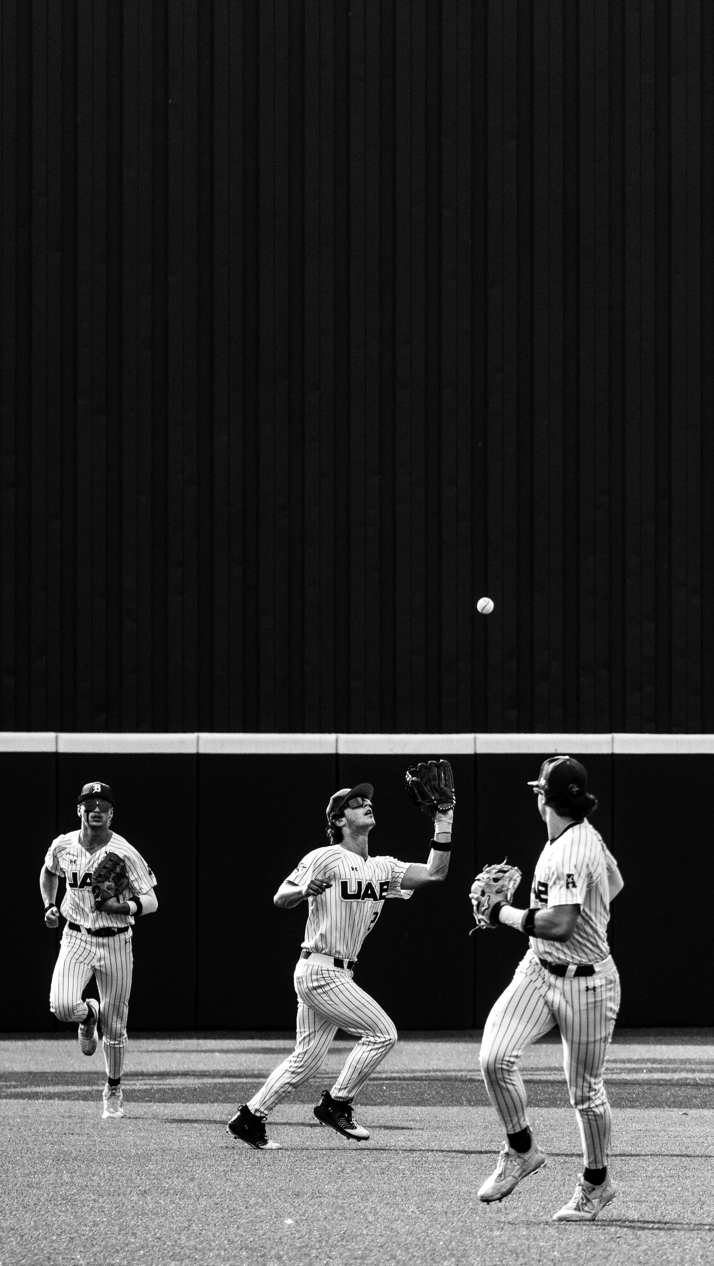Black-and-white photo of three baseball players on the field, with two players facing each other and one running in the background, all wearing striped uniforms with "UAB" on the front, and a baseball in mid-air above them.
