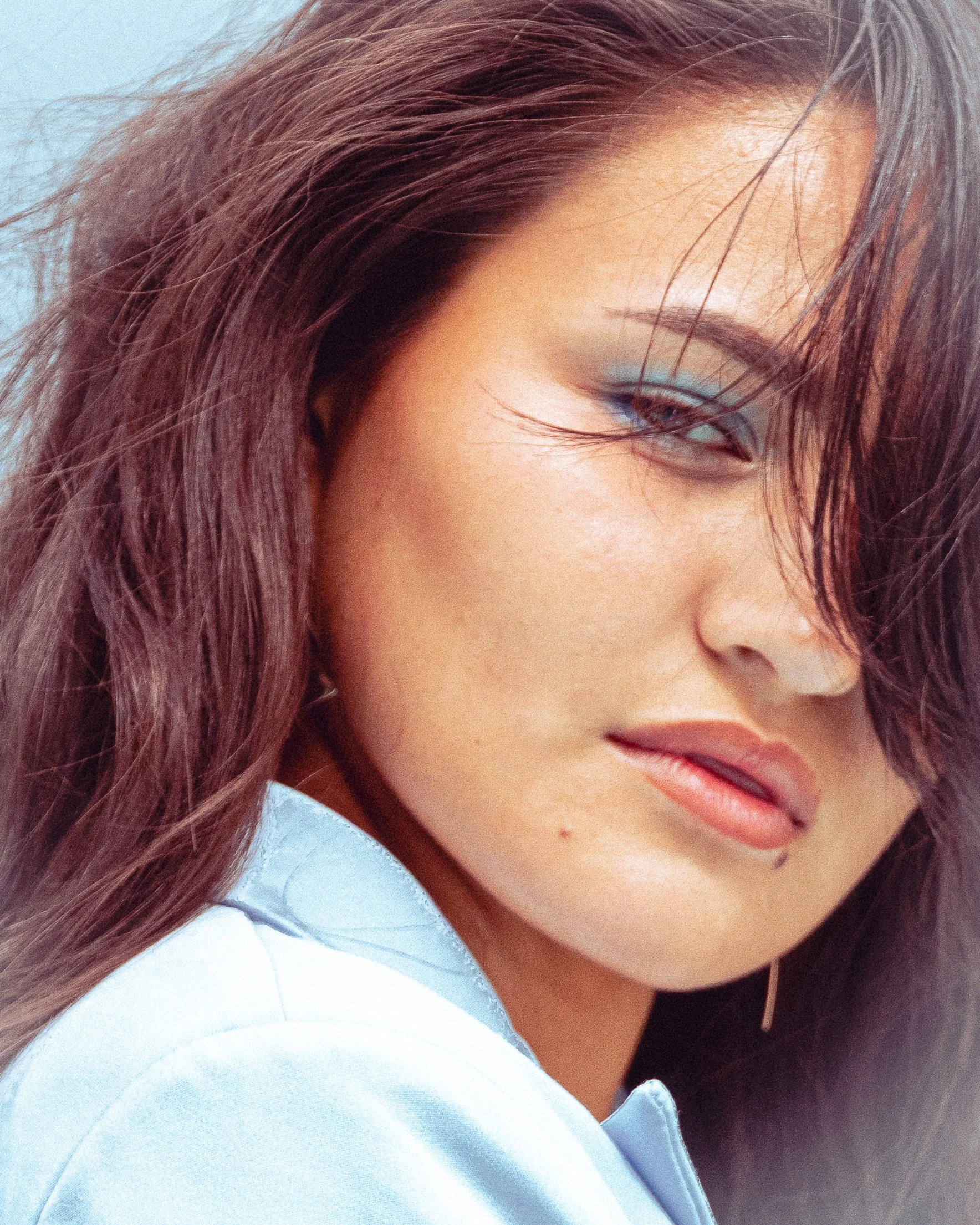 Close-up portrait of a woman with wavy brown hair, wearing light blue eyeshadow and a light blue shirt.