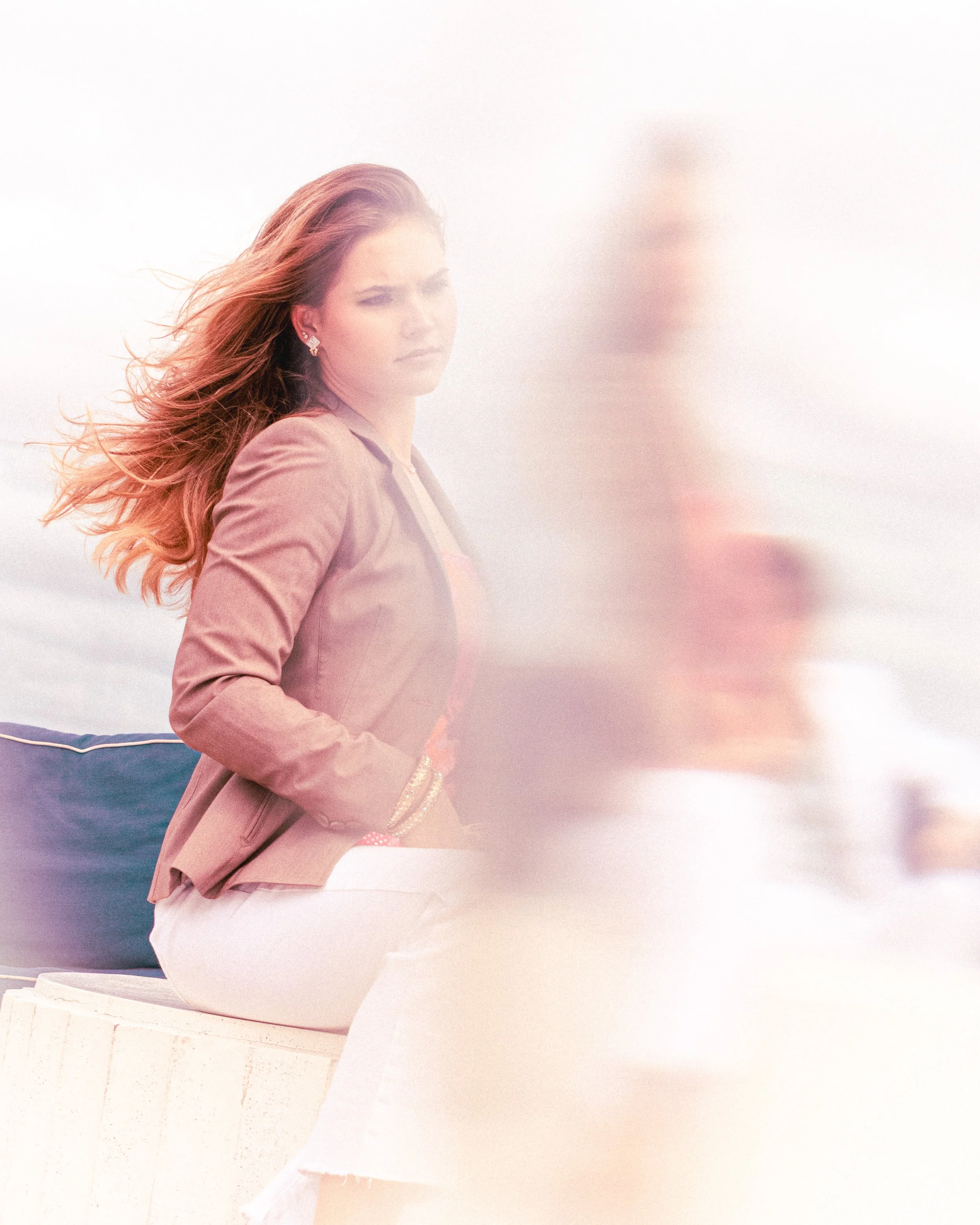 Woman with long, wavy hair wearing a pink jacket and white pants, sitting outdoors with cloudy sky background, partially obscured by a blurred figure in the right foreground.