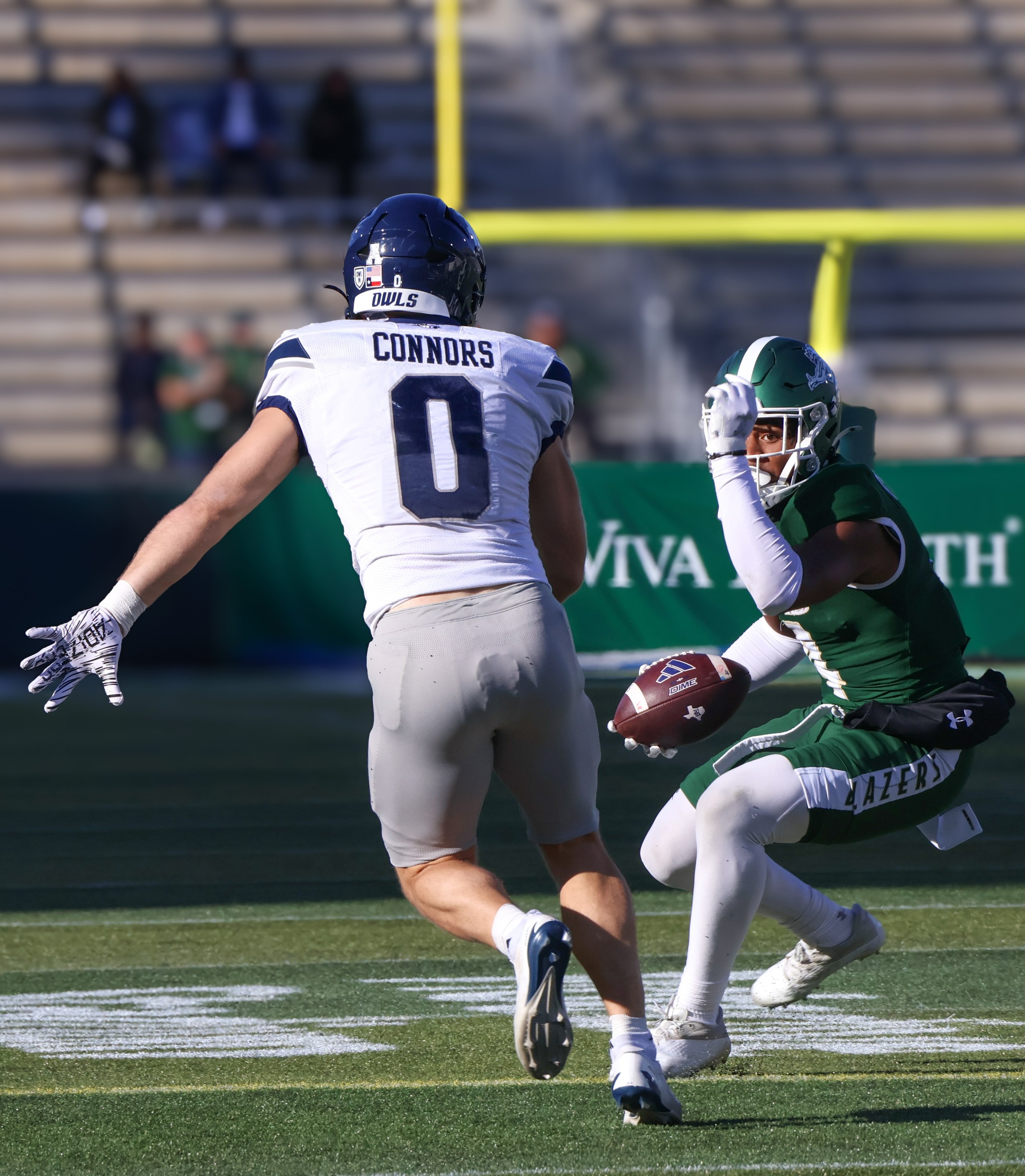 An American football game in progress with a player in a green uniform holding the ball and a player in a white uniform attempting to tackle him.