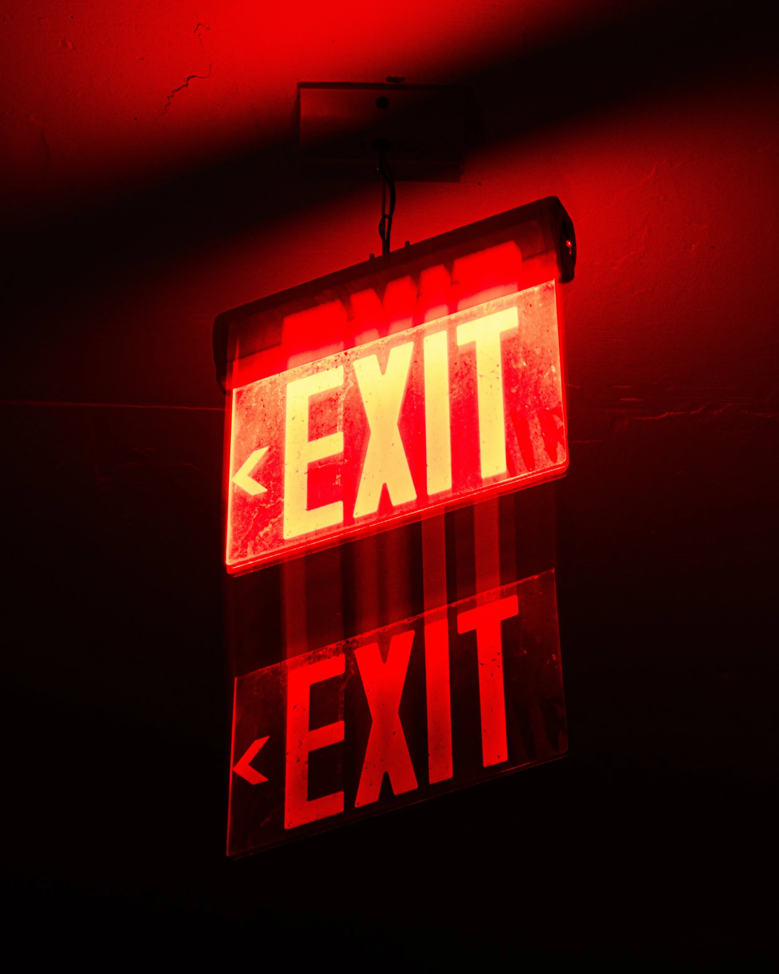 Red illuminated exit sign with arrows pointing to the left, mounted on a ceiling in a dark setting.