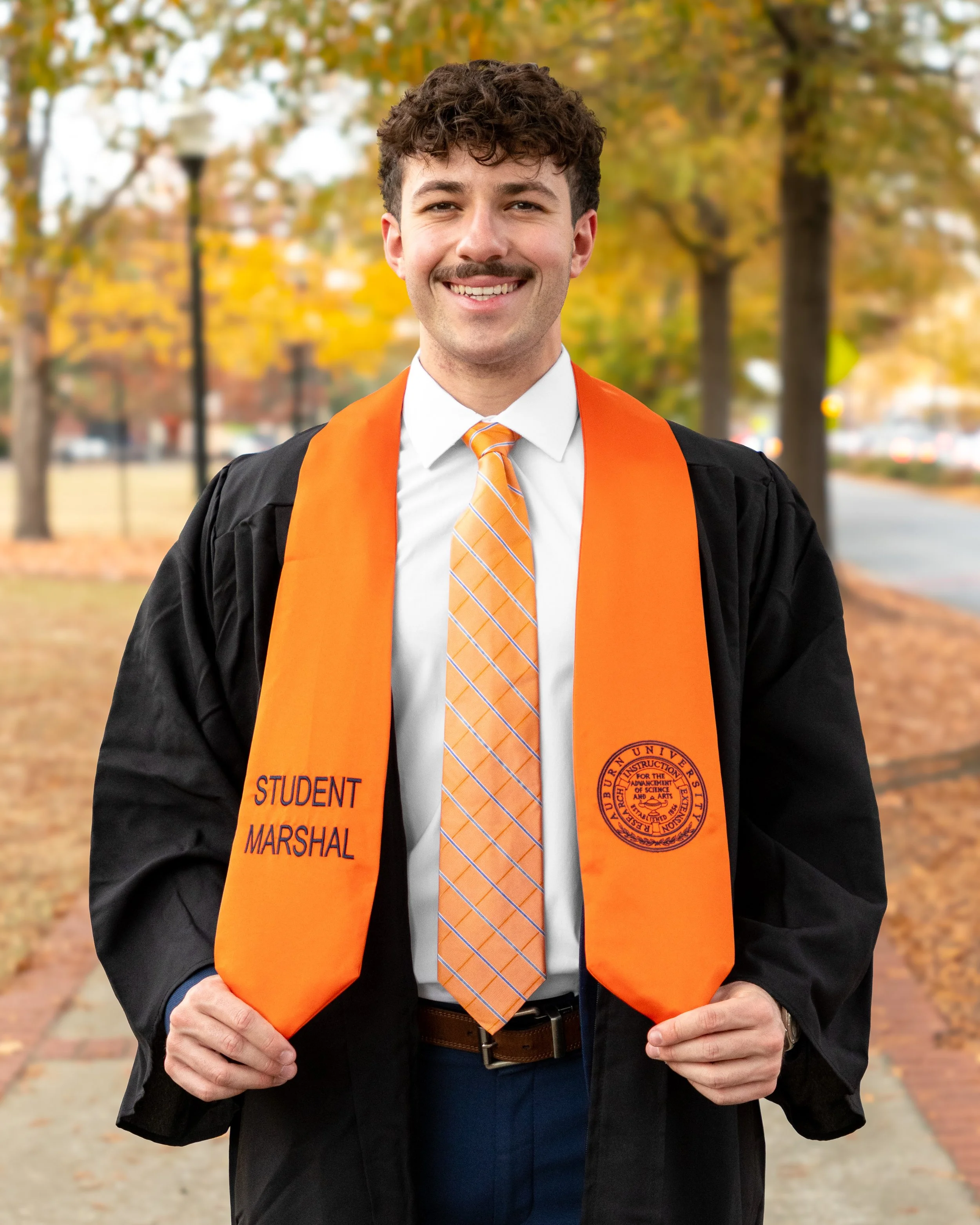 Young man in graduation gown and cap, holding an orange sash with 'Student Marshal' and university seal, smiling outdoors with fall foliage in the background.