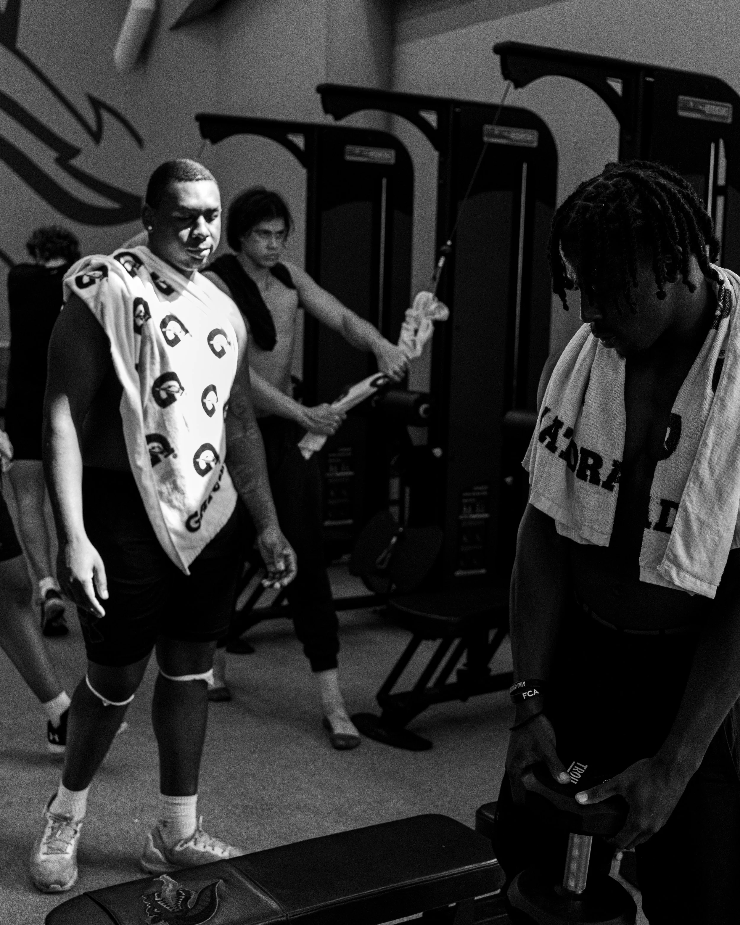 A black and white photo of athletes and trainers in a gym, with one athlete holding a dumbbell and sweat towels around necks.