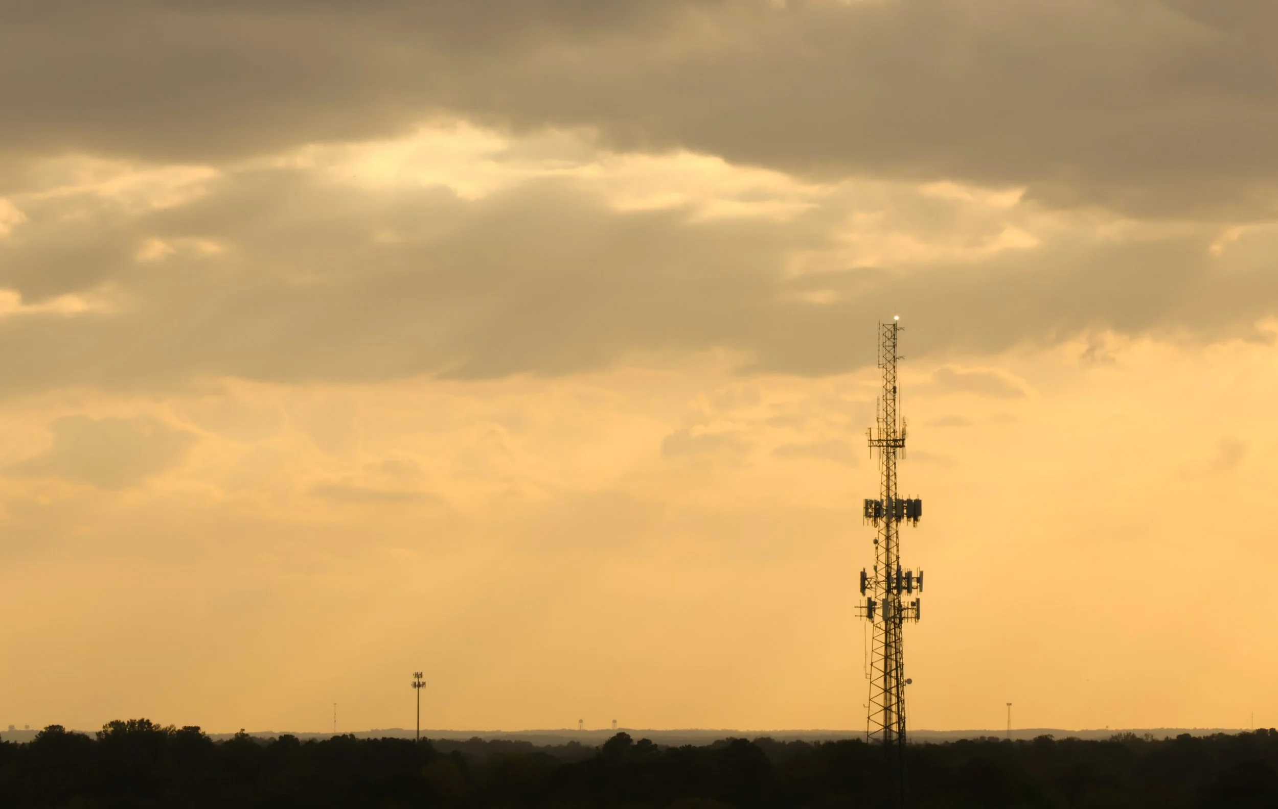 A tall radio tower with multiple antennas stands against a cloudy sky during sunset, with a few other smaller towers visible on the horizon.