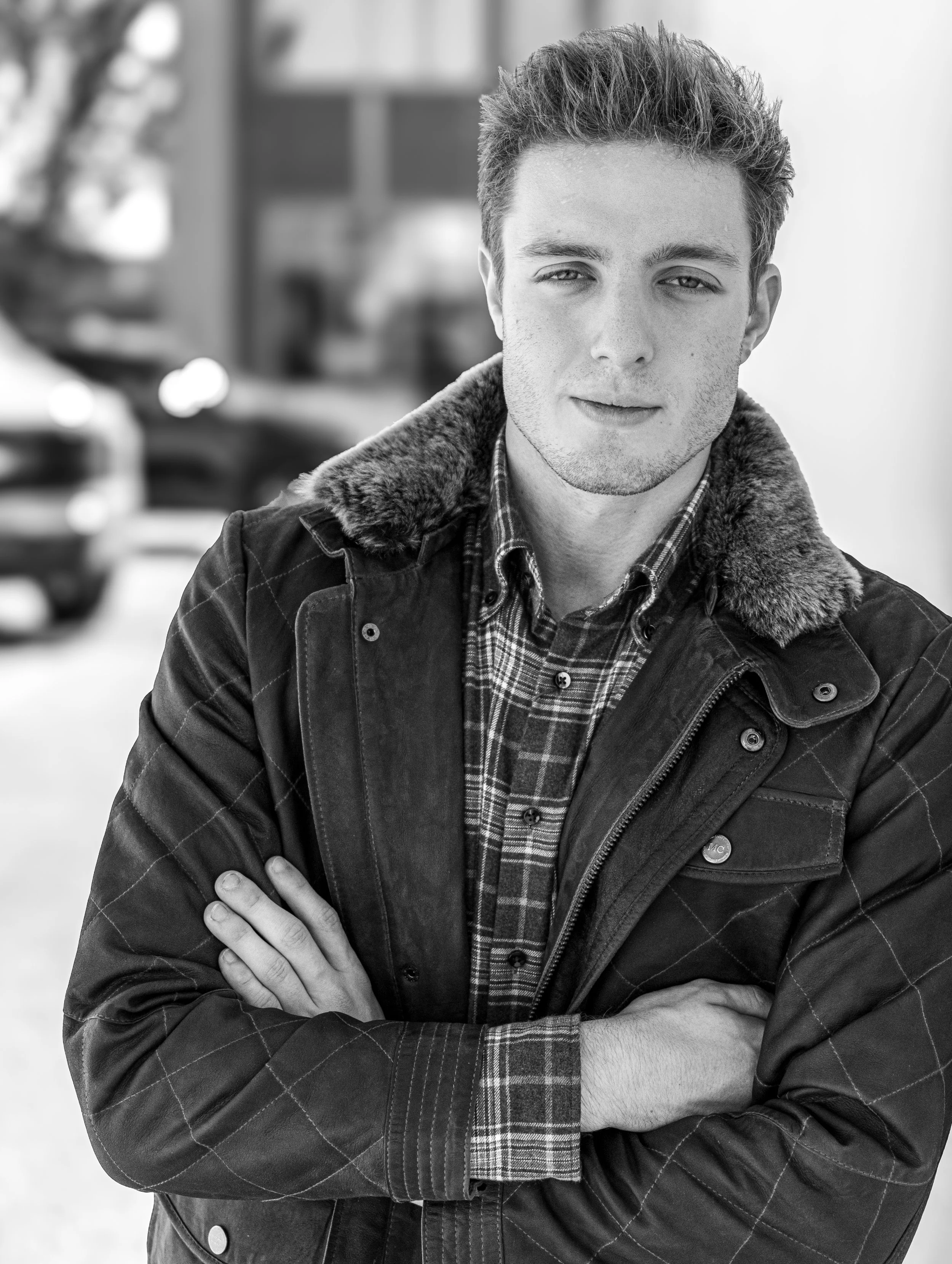 Black and white photo of a young man with crossed arms, wearing a plaid shirt and a jacket with a fur collar, standing outdoors.