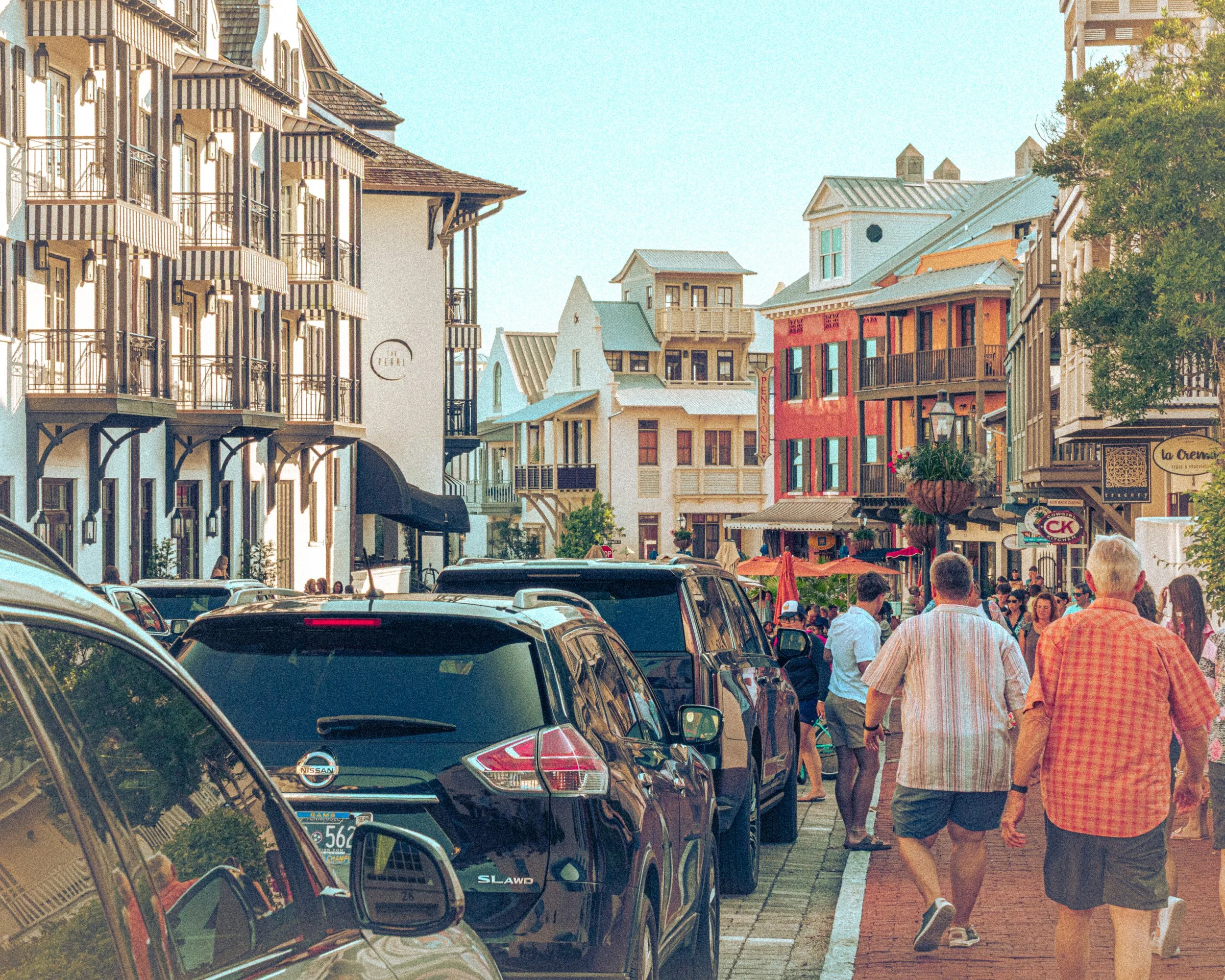 A busy street scene in a charming town with colorful buildings, parked cars, and pedestrians walking on the sidewalk, some wearing casual summer clothes.