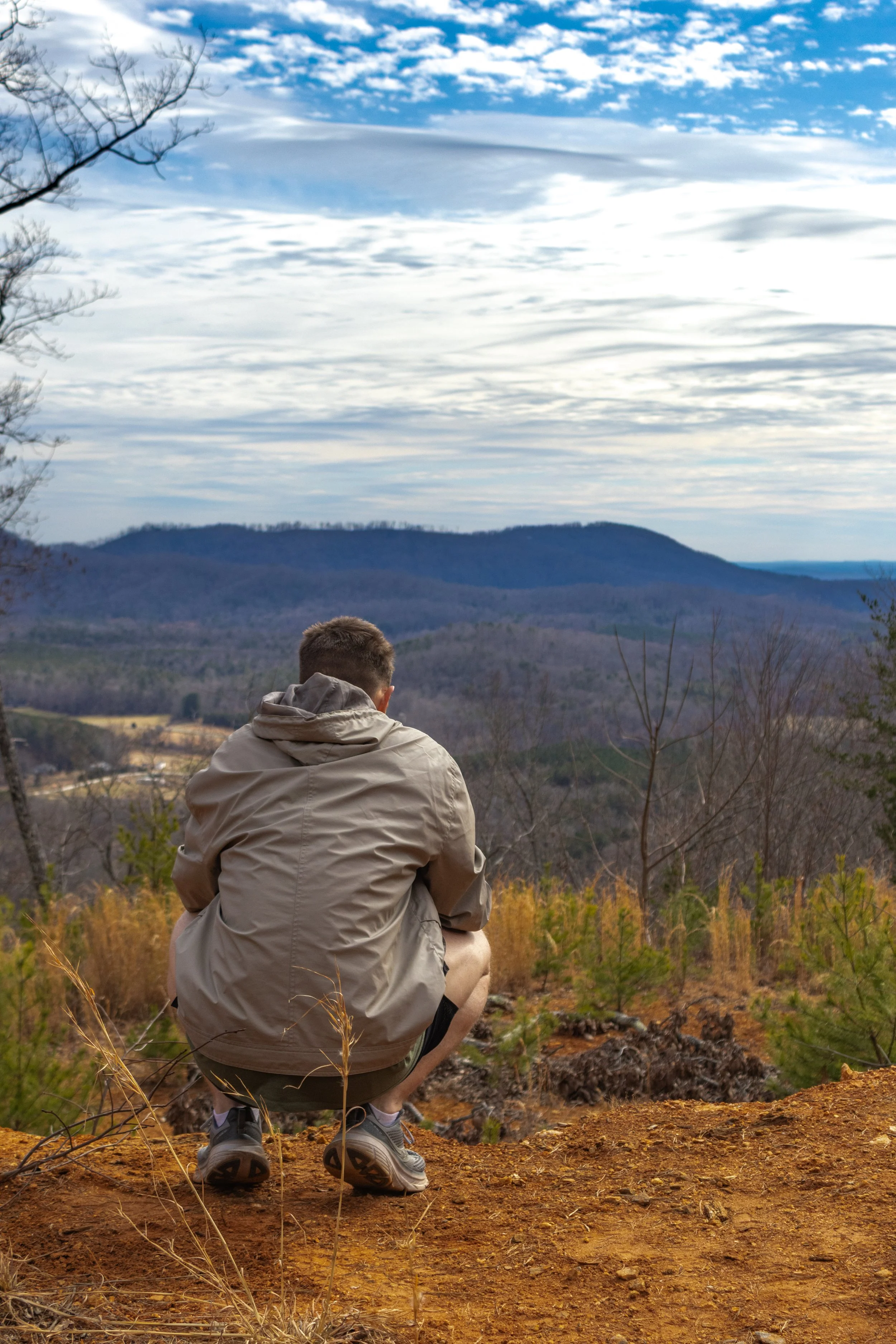 A person squatting on a dirt trail overlooking a landscape of rolling hills and mountains with a cloudy sky overhead.