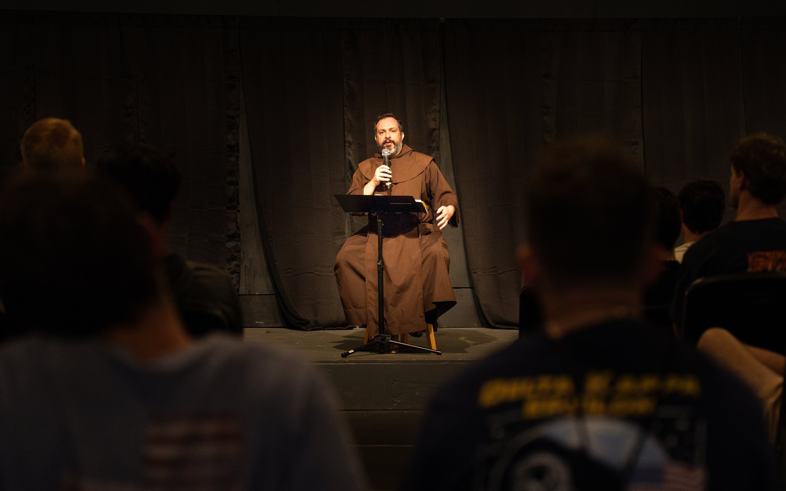 A man dressed in a brown robe speaking into a microphone on stage in front of an audience, with a dark curtain backdrop.