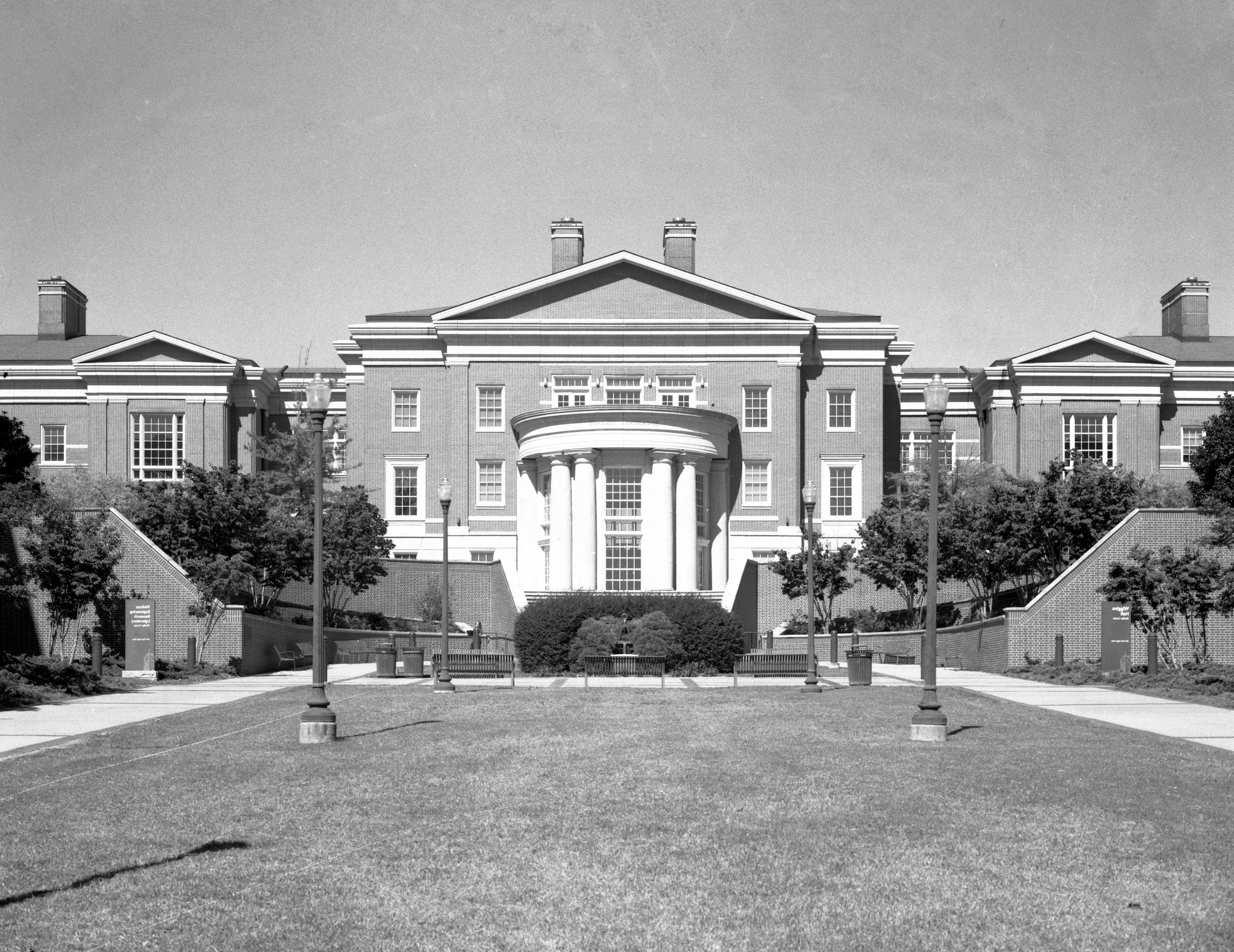 A large, three-story brick building with classical architectural details, including columns and a central curved entrance, surrounded by trees, lampposts, and a well-maintained lawn.