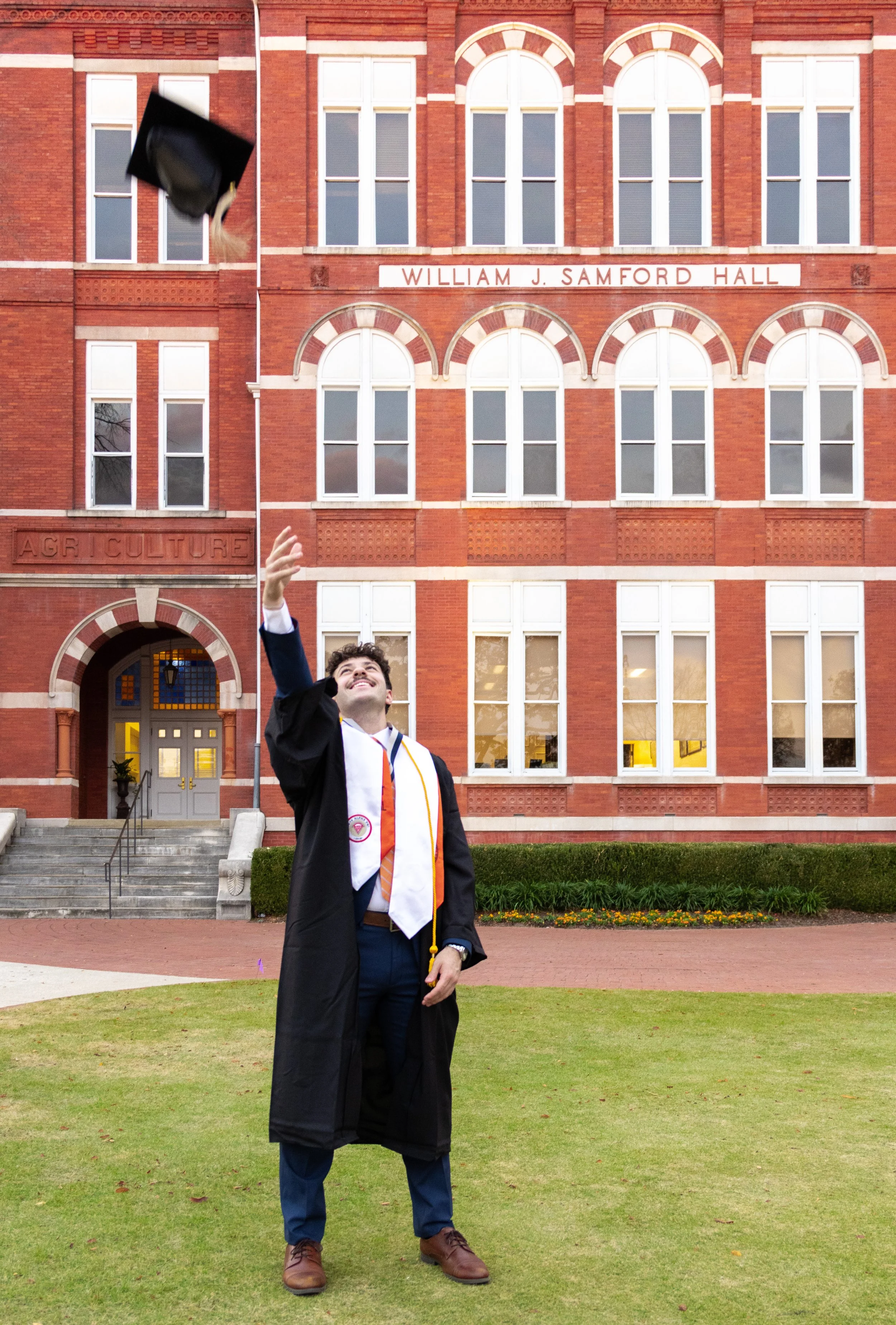 A young man in a graduation gown and cap celebrating outside William J. Samford Hall at Auburn University, tossing his cap in the air.
