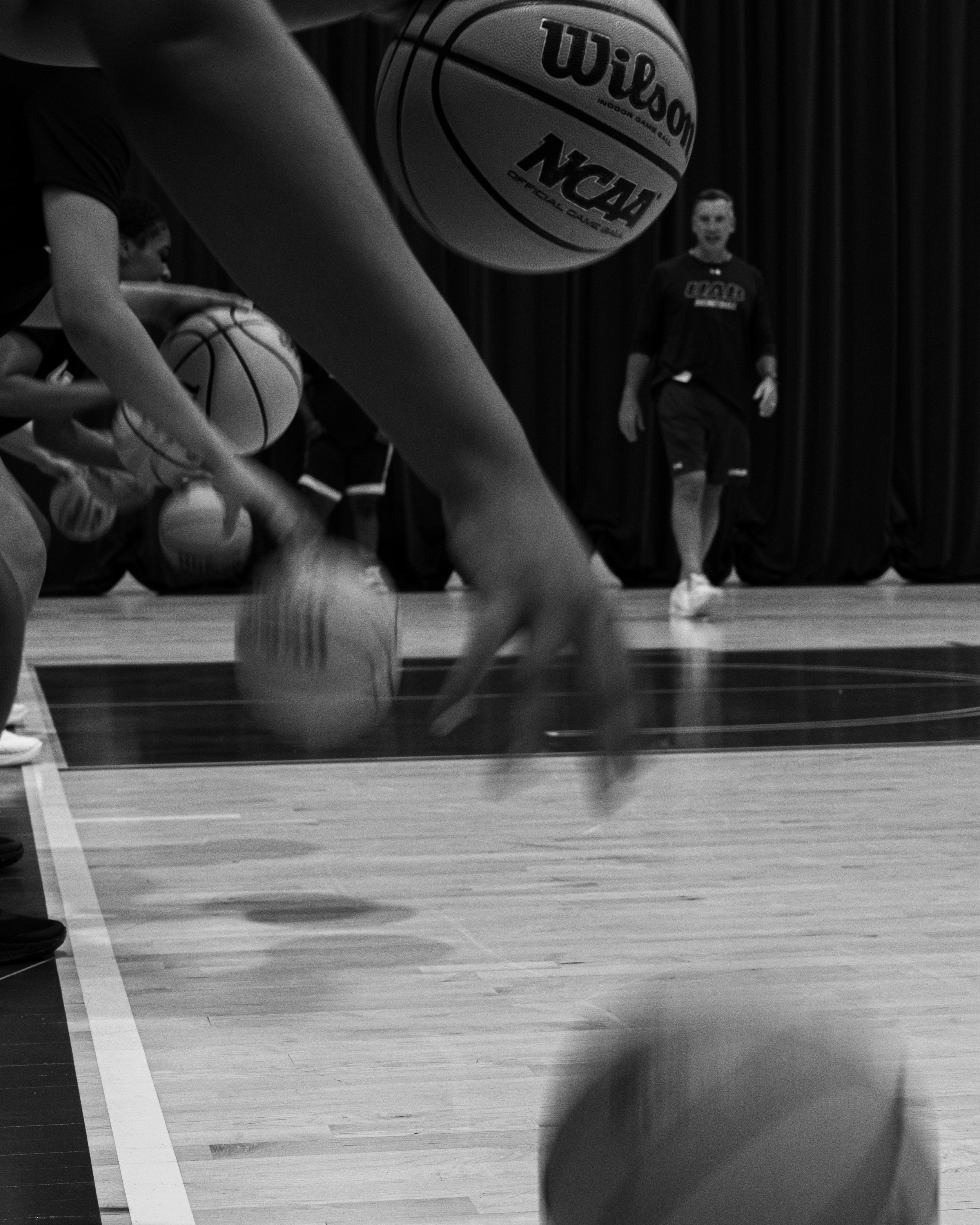 Black and white photo of a basketball practice session with players dribbling basketballs and a coach observing.