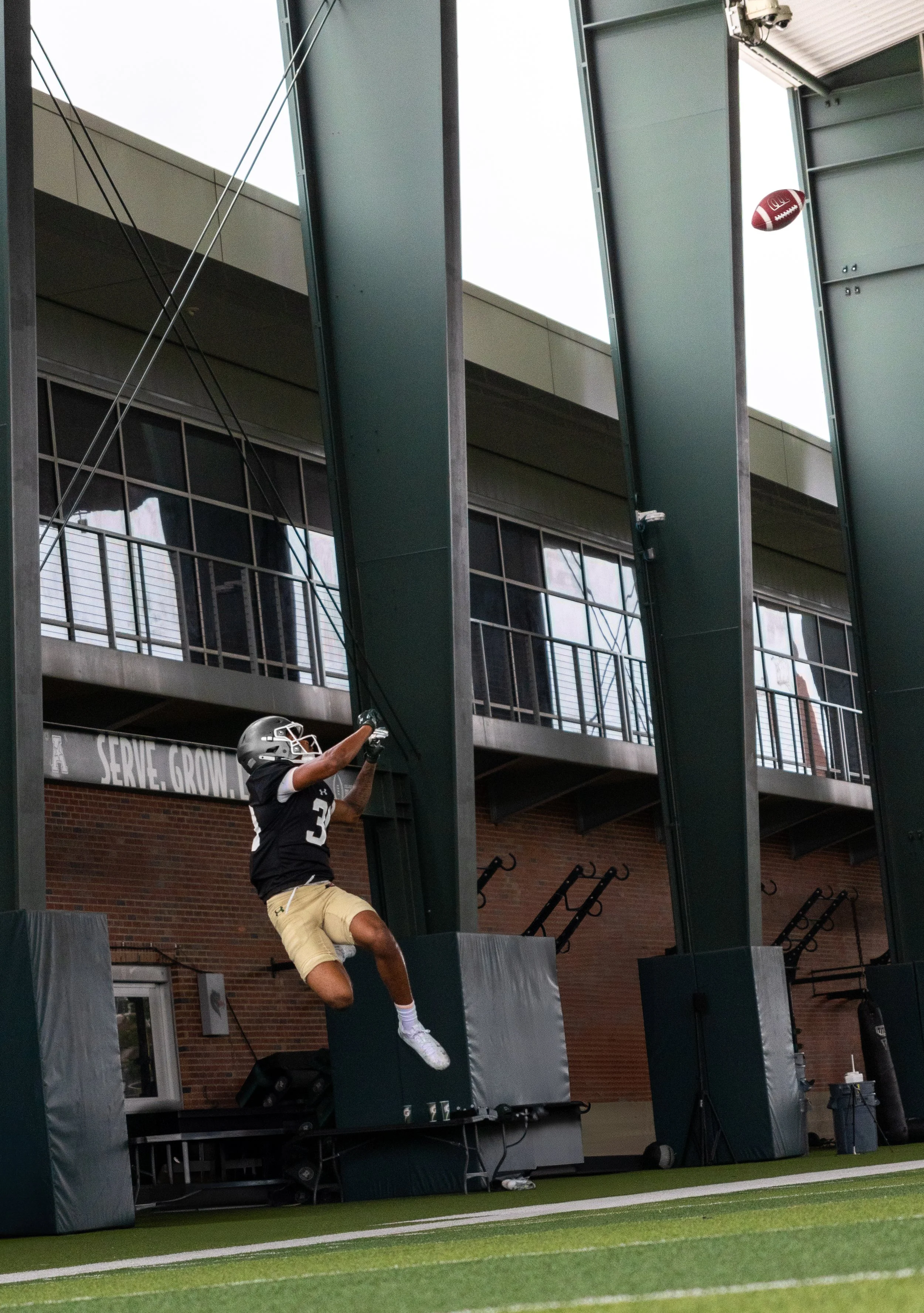 Football player in a black jersey and beige shorts jumps to catch a football at a stadium with green turf, modern architectural structures, and a brick building in the background.