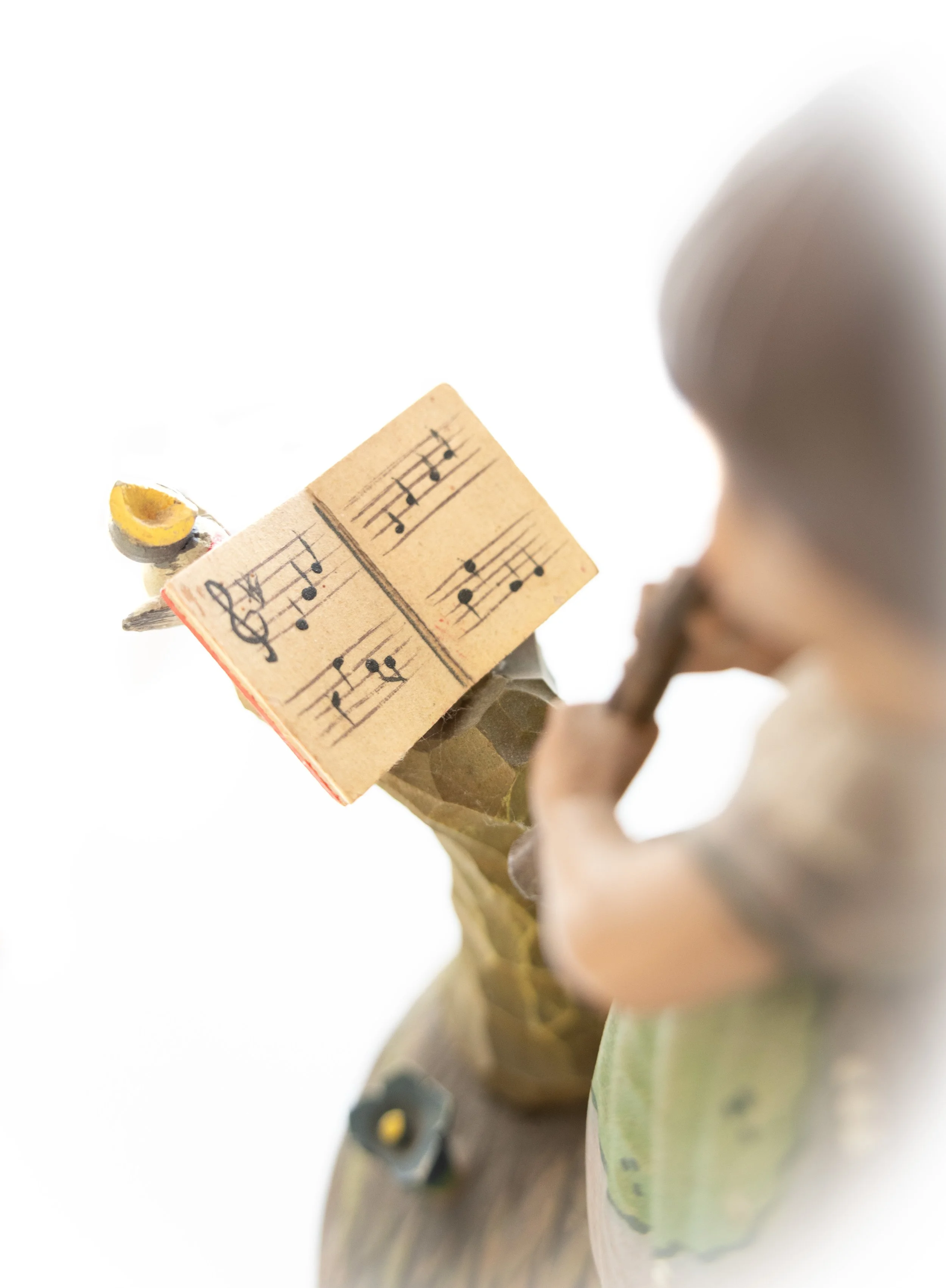 Close-up of a figurine of a child playing a flute and holding sheet music, with a small yellow bird perched near the sheet music.