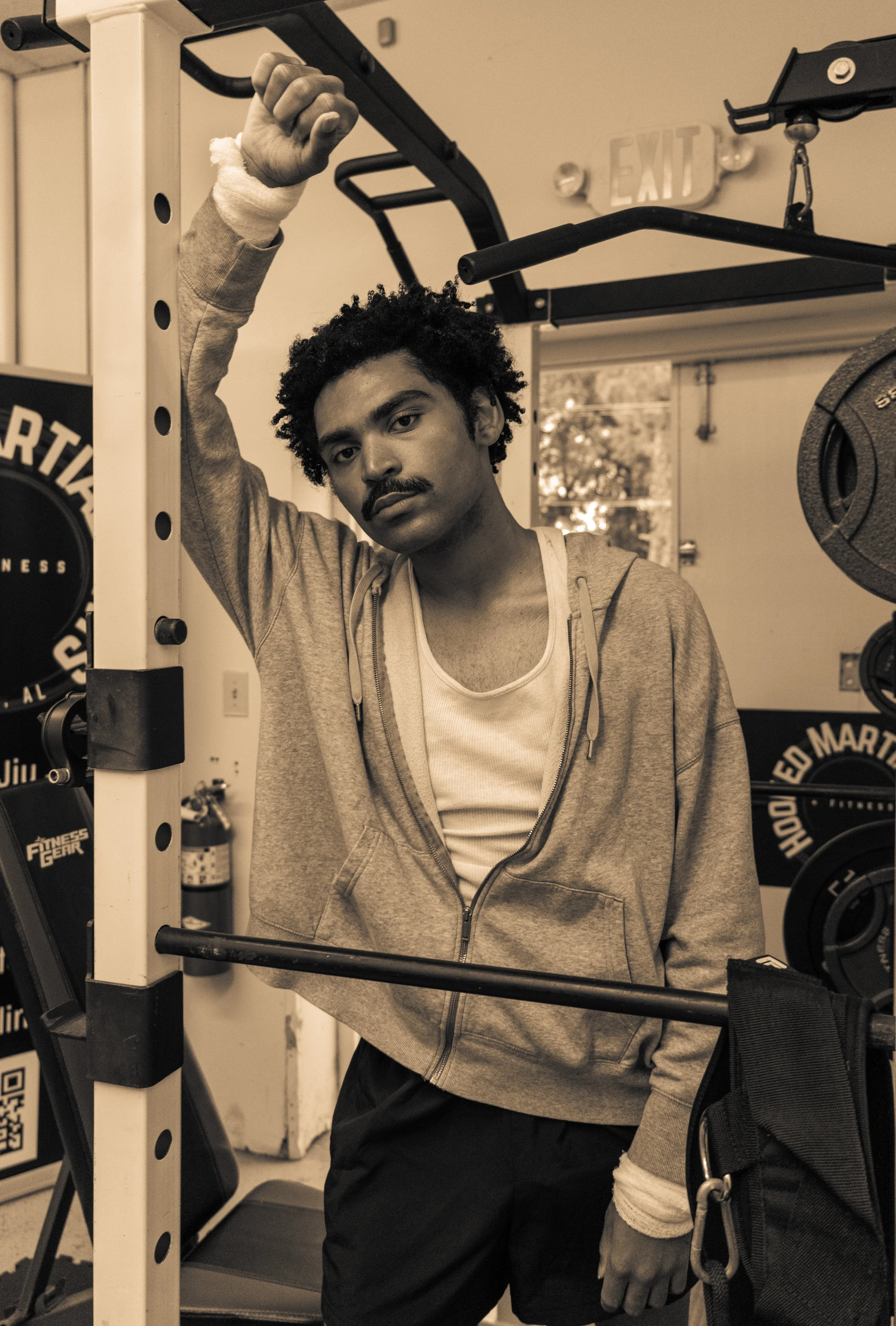 A young man with curly hair and a mustache leaning against gym equipment, wearing a hoodie and a white tank top, in a gym setting.