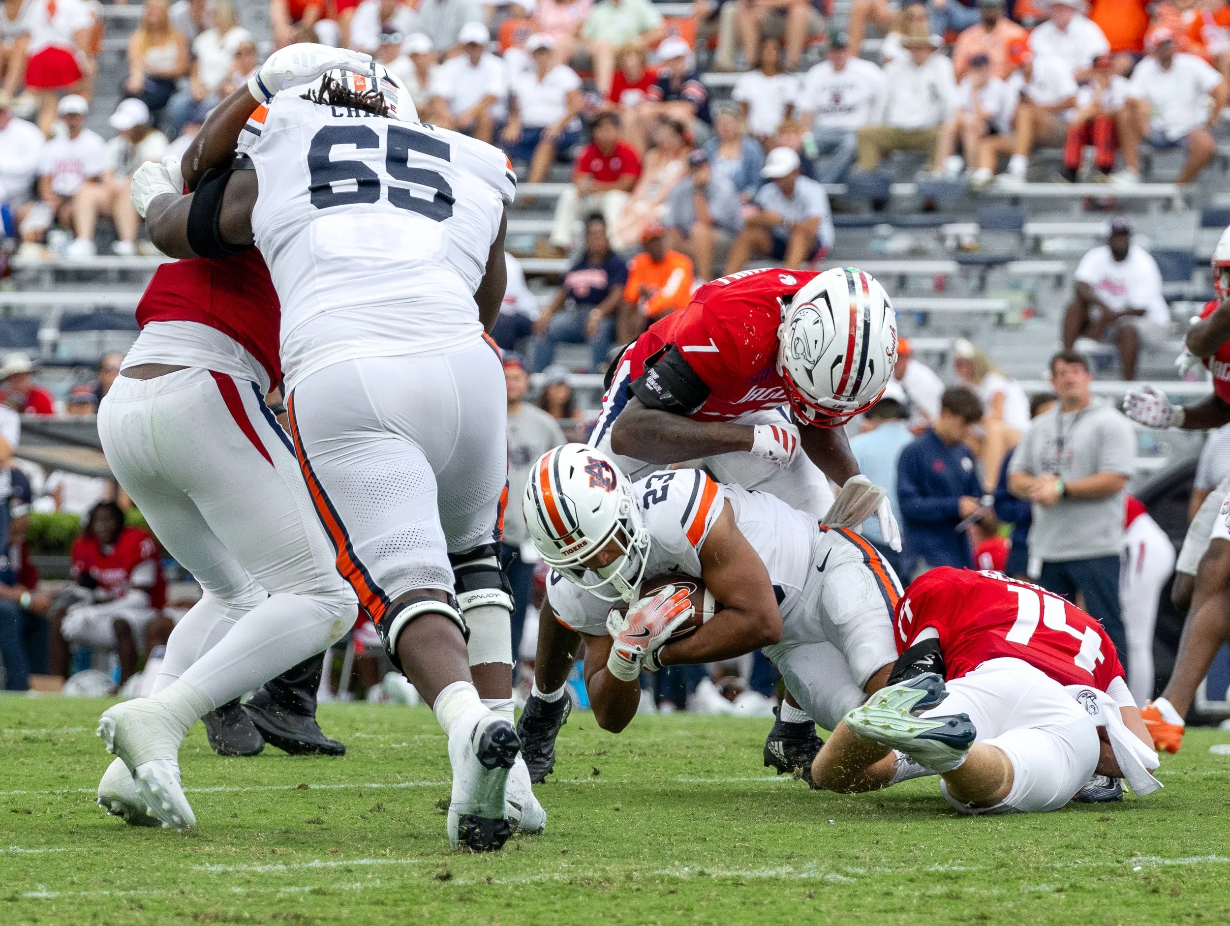 An American football game scene with players tackling an opponent on the field, a crowd of spectators watching from the stands.