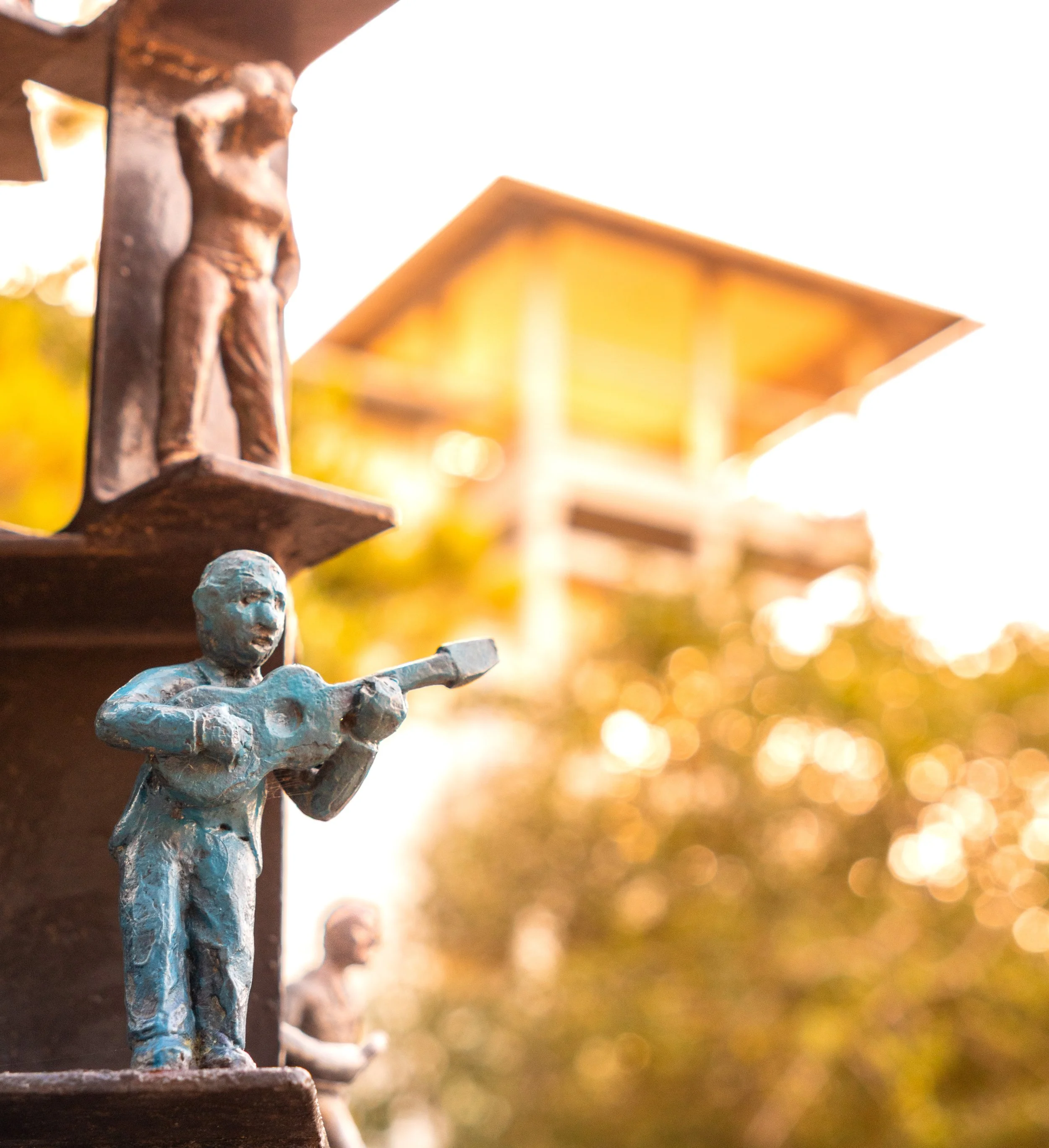 Close-up of small, colorful figurines on a sculpture with a blurred background of trees and a structure under warm sunlight.