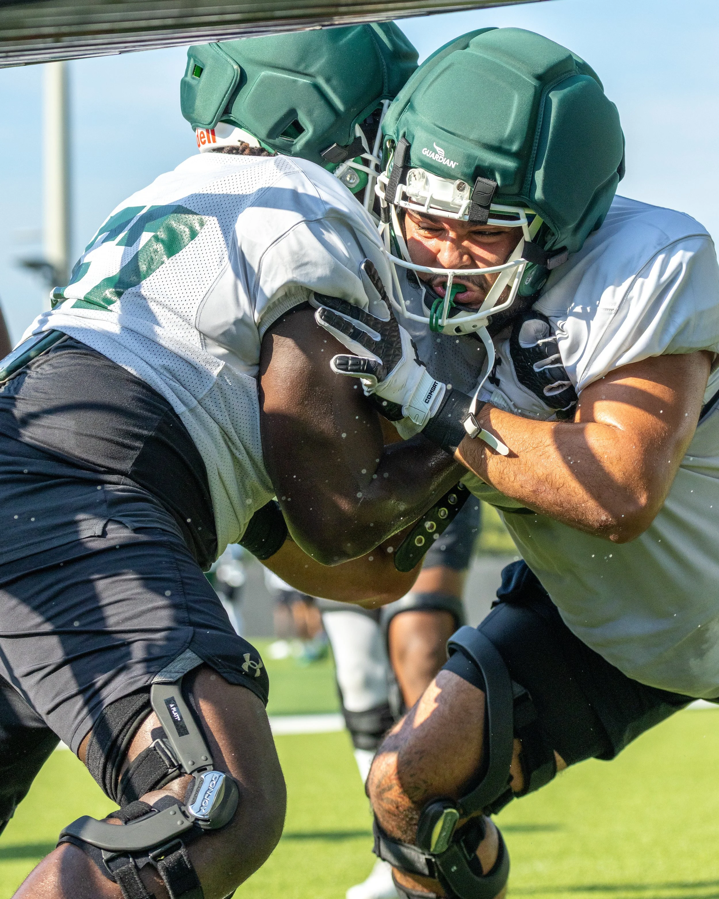 Two football players wearing helmets and gear engage in a blocking drill during practice on a grassy field.