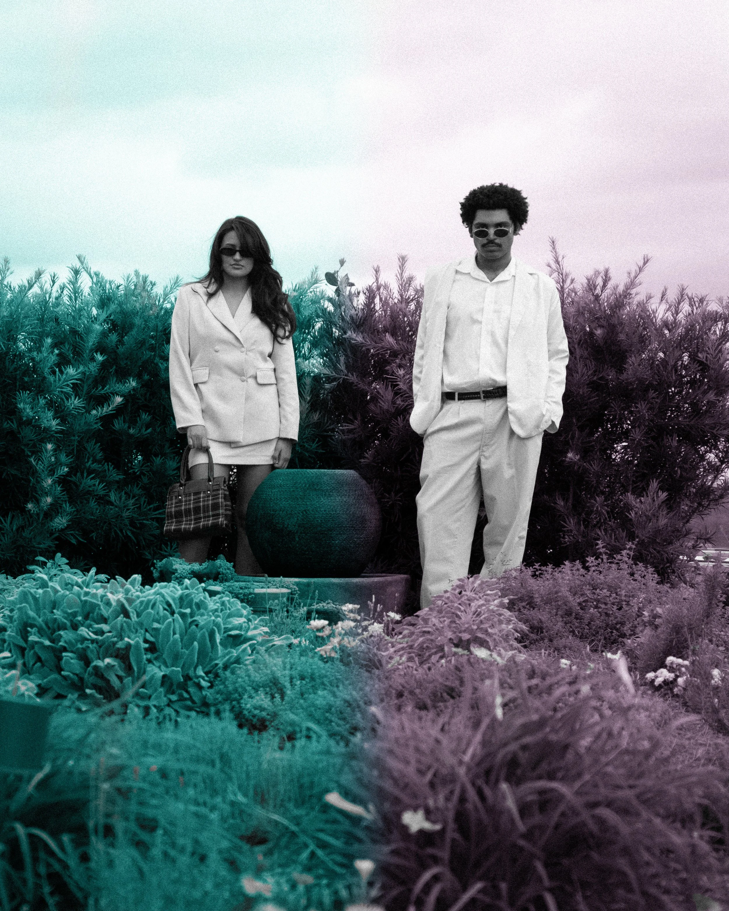 Two people wearing white outfits and sunglasses standing among lush plants and shrubs outdoors.
