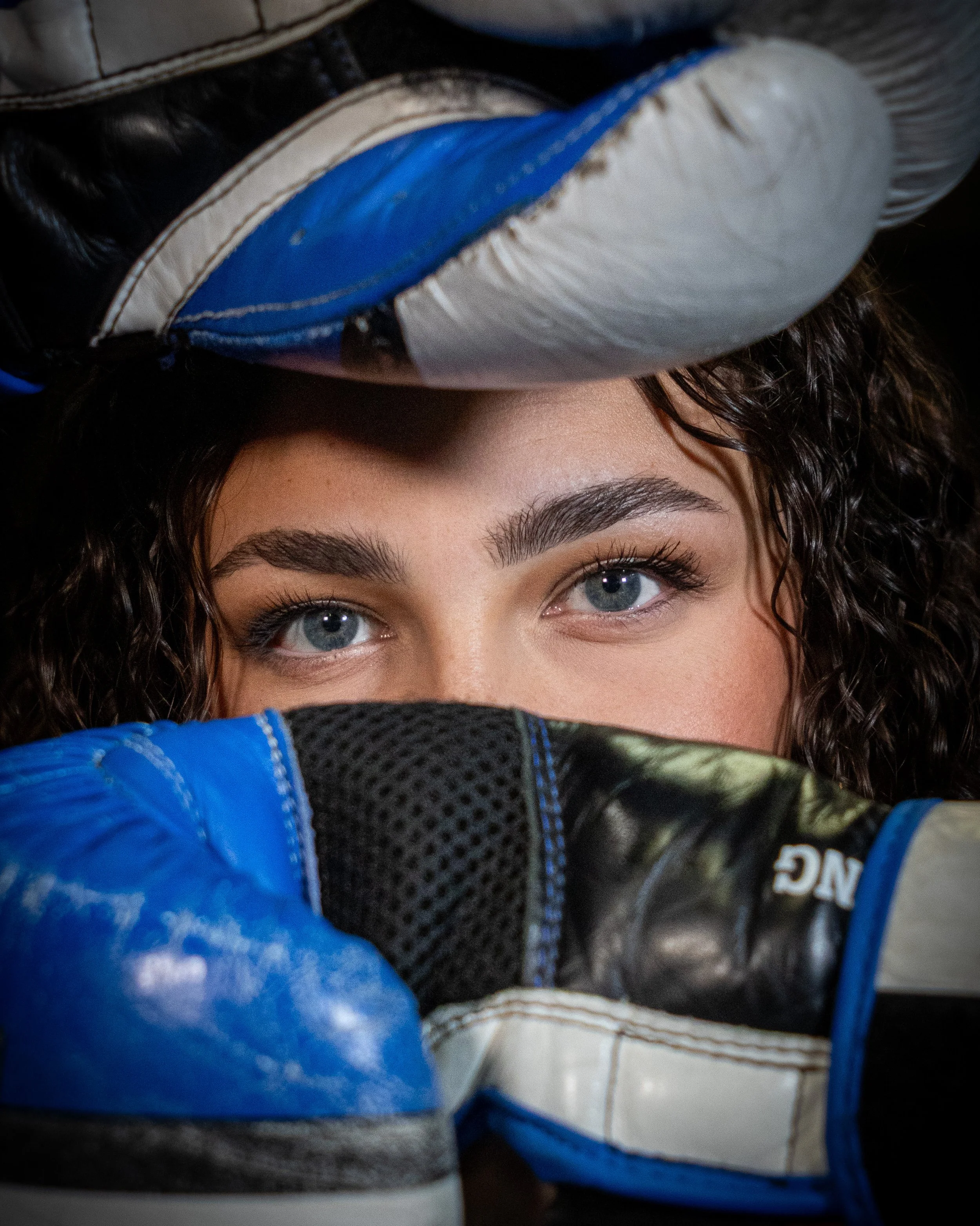 Close-up of a woman with curly hair and blue eyes hiding her face behind her arms, wearing boxing gloves.