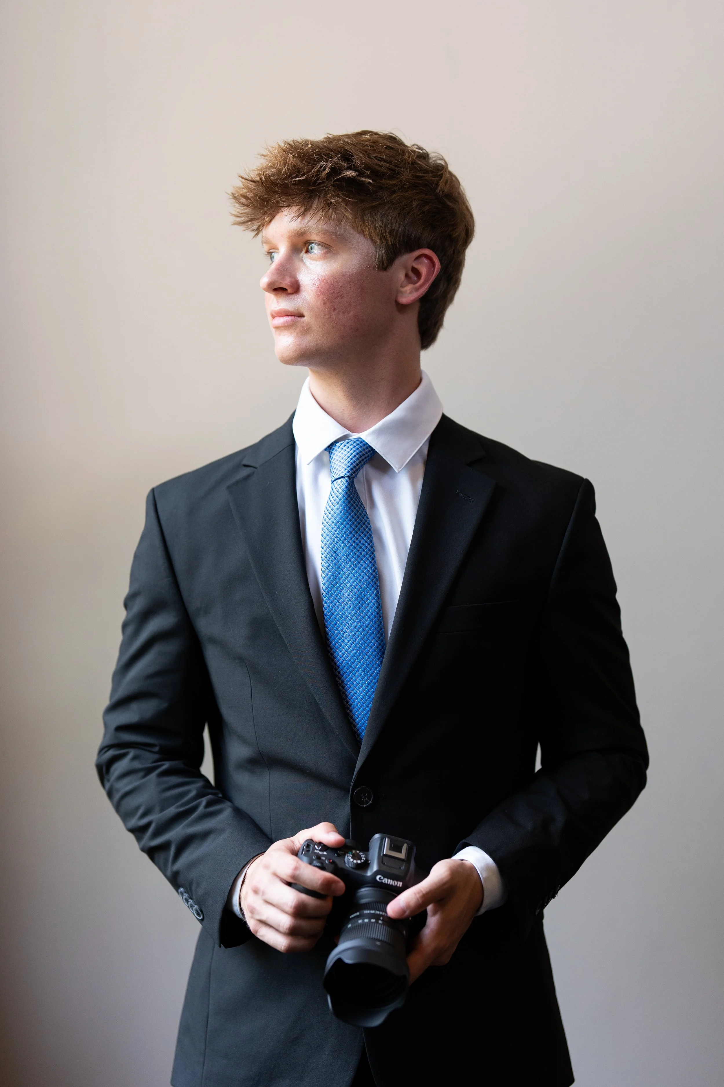 A young man in a black suit and blue tie holding a Canon camera, looking to the left.
