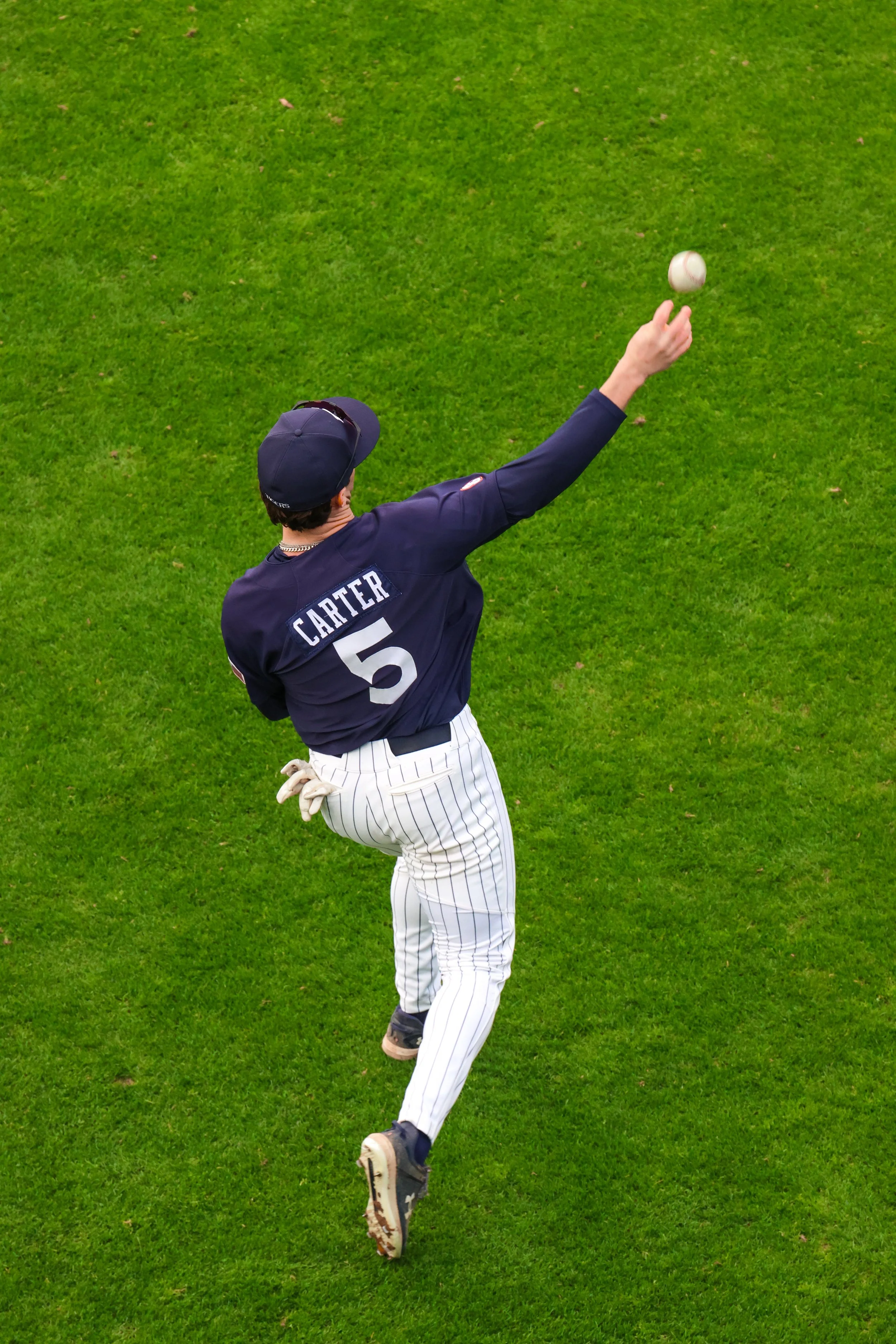 A baseball player in a dark blue jacket with the number 5 and the name Carter, white striped pants, and a dark cap, throws a baseball on a green field.