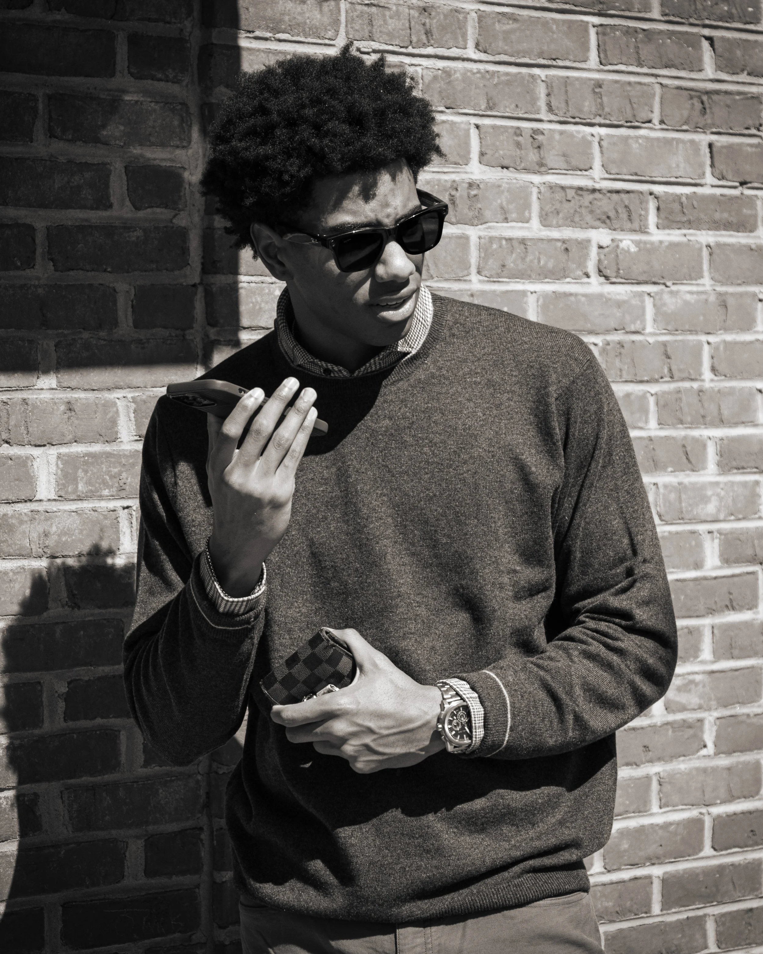 A young man with curly hair, wearing sunglasses, a sweater, and a watch, standing against a brick wall, holding a phone and a wallet.