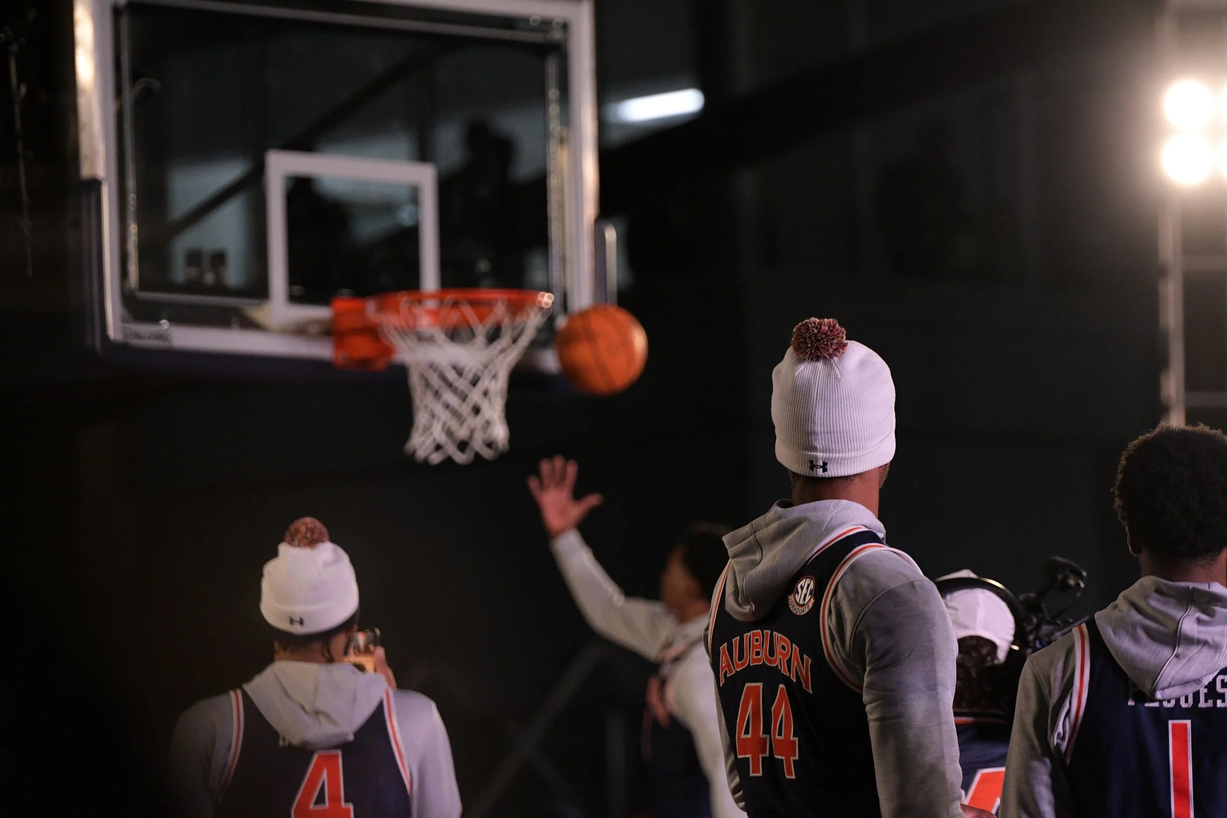People in Auburn basketball jerseys watching a basketball shot into the hoop, with a basketball in midair near the hoop.